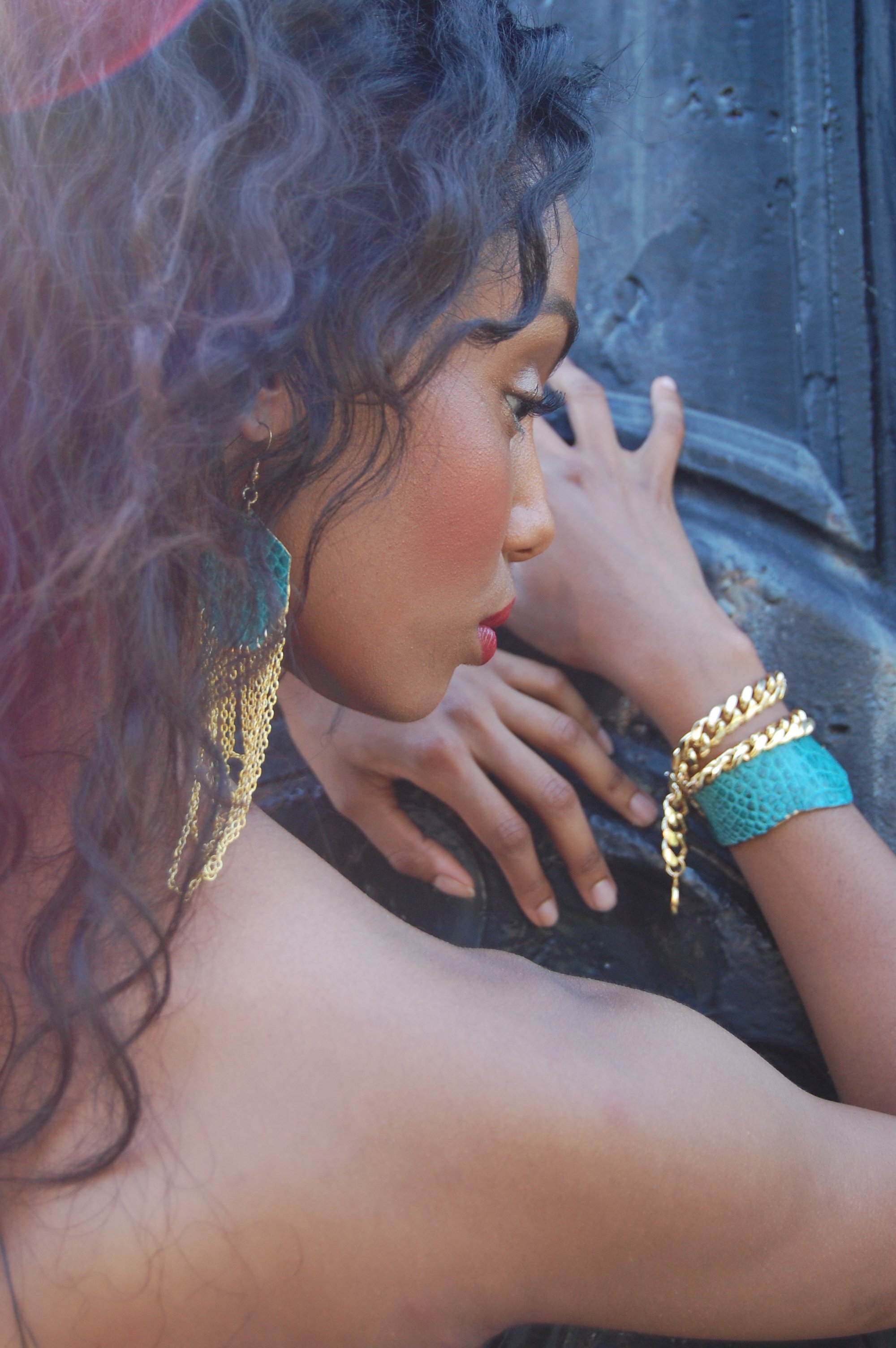 A woman with curly dark hair and gold jewelry, resting against a textured dark surface, with makeup including blush and red lipstick. Holy Harlot Jewelry photographed by artist Magda Parasidis, fashion photographer.