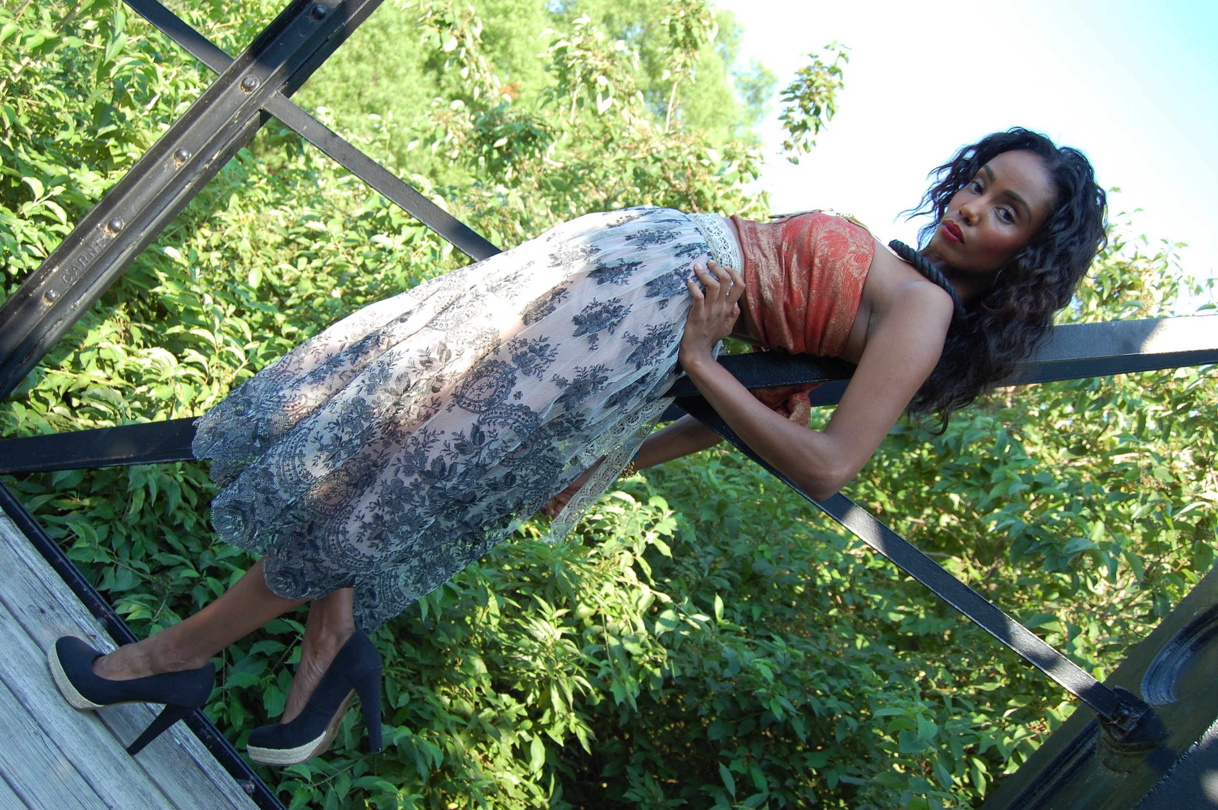 A woman with curly black hair and dark skin wearing a floral skirt, black high heels, and a reddish-orange top posing on a black metal structure surrounded by green foliage.