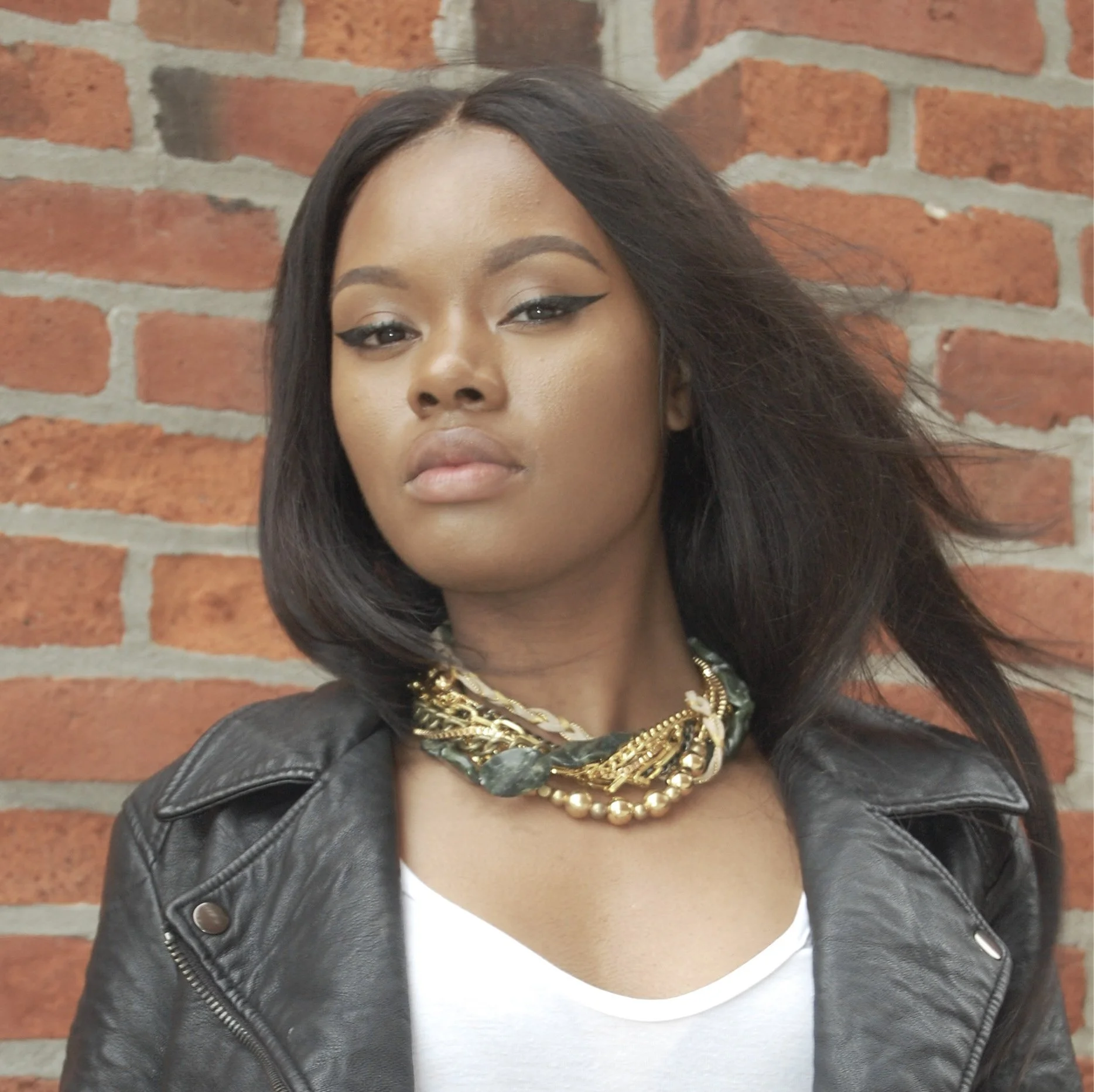 A woman with shoulder-length black hair, wearing makeup with winged eyeliner, a chunky gold and green necklace, a white top, and a black leather jacket, standing in front of a brick wall.