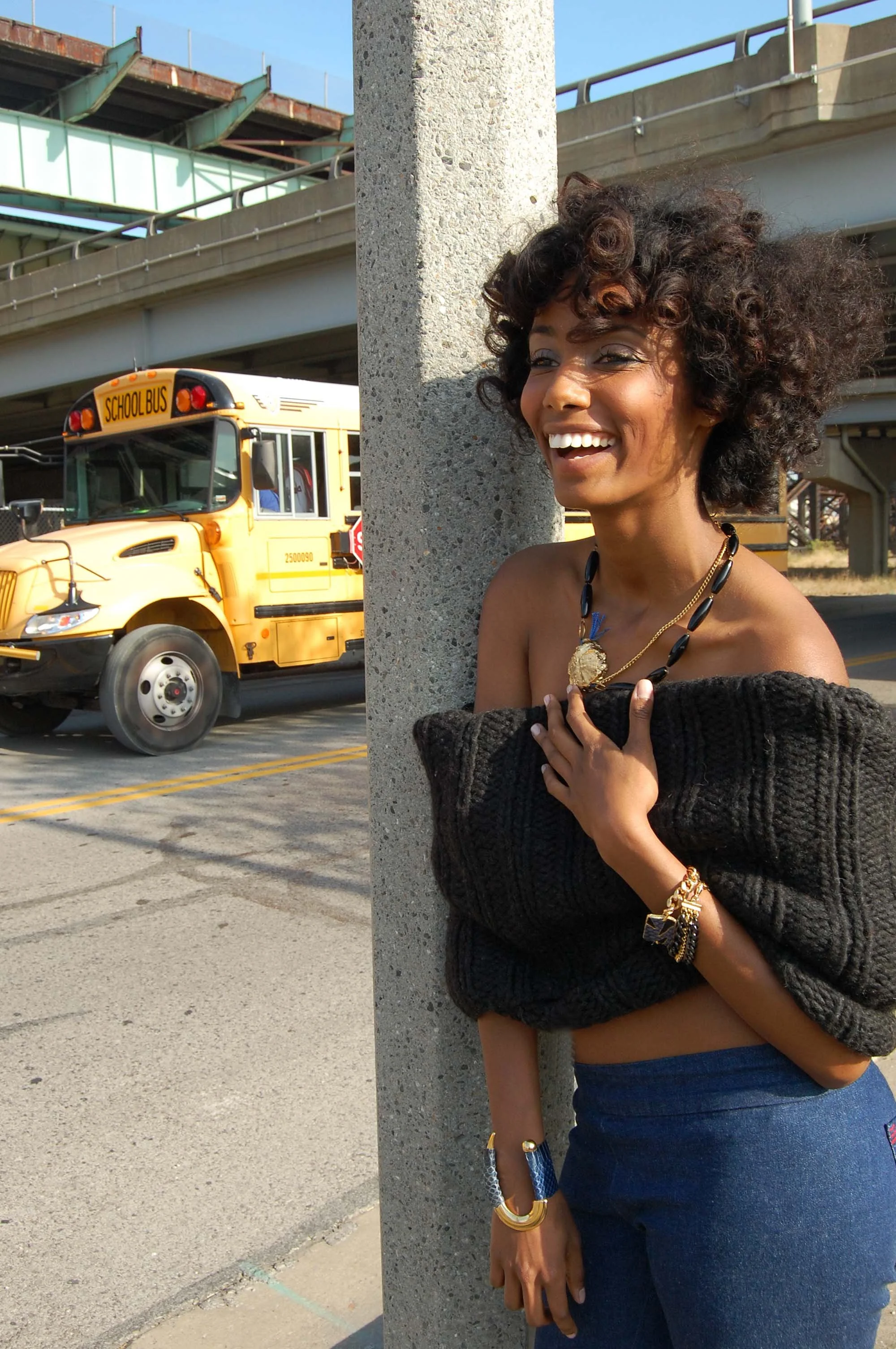 A woman with curly hair smiling and holding her chest, standing beside a concrete pole on a city street with a yellow school bus in the background.