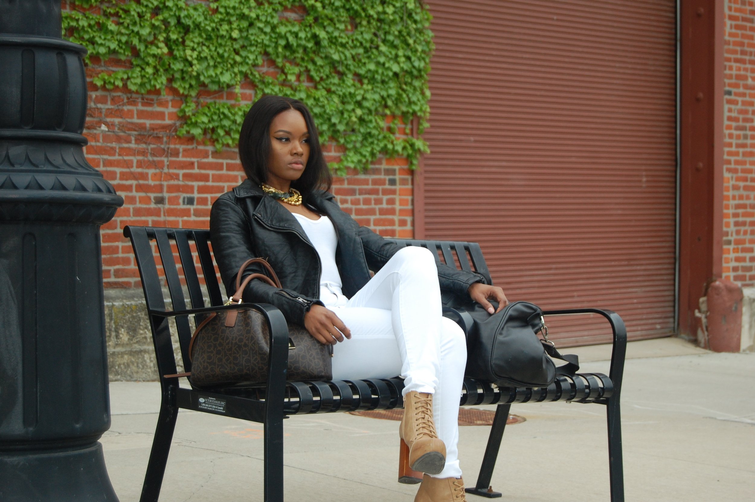 A woman sitting alone on a black metal bench outdoors, dressed in white pants, a white top, a black leather jacket, and tan lace-up boots, with handbags placed on the bench beside her, against a background of a brick wall with green ivy and a red garage door.