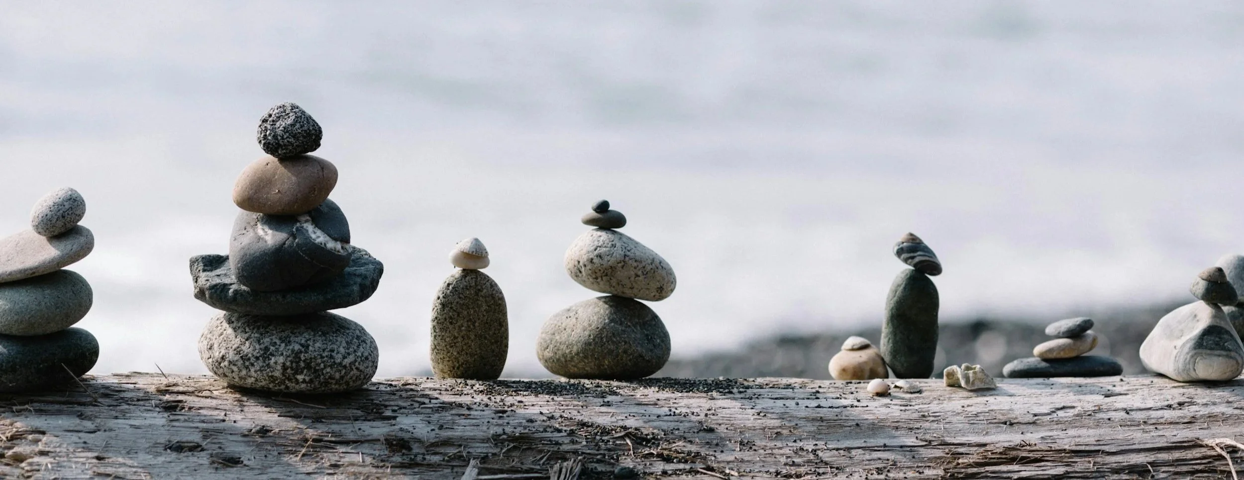Multiple stacks of rocks arranged on a weathered wooden surface near the water, with a blurred ocean or lake in the background.