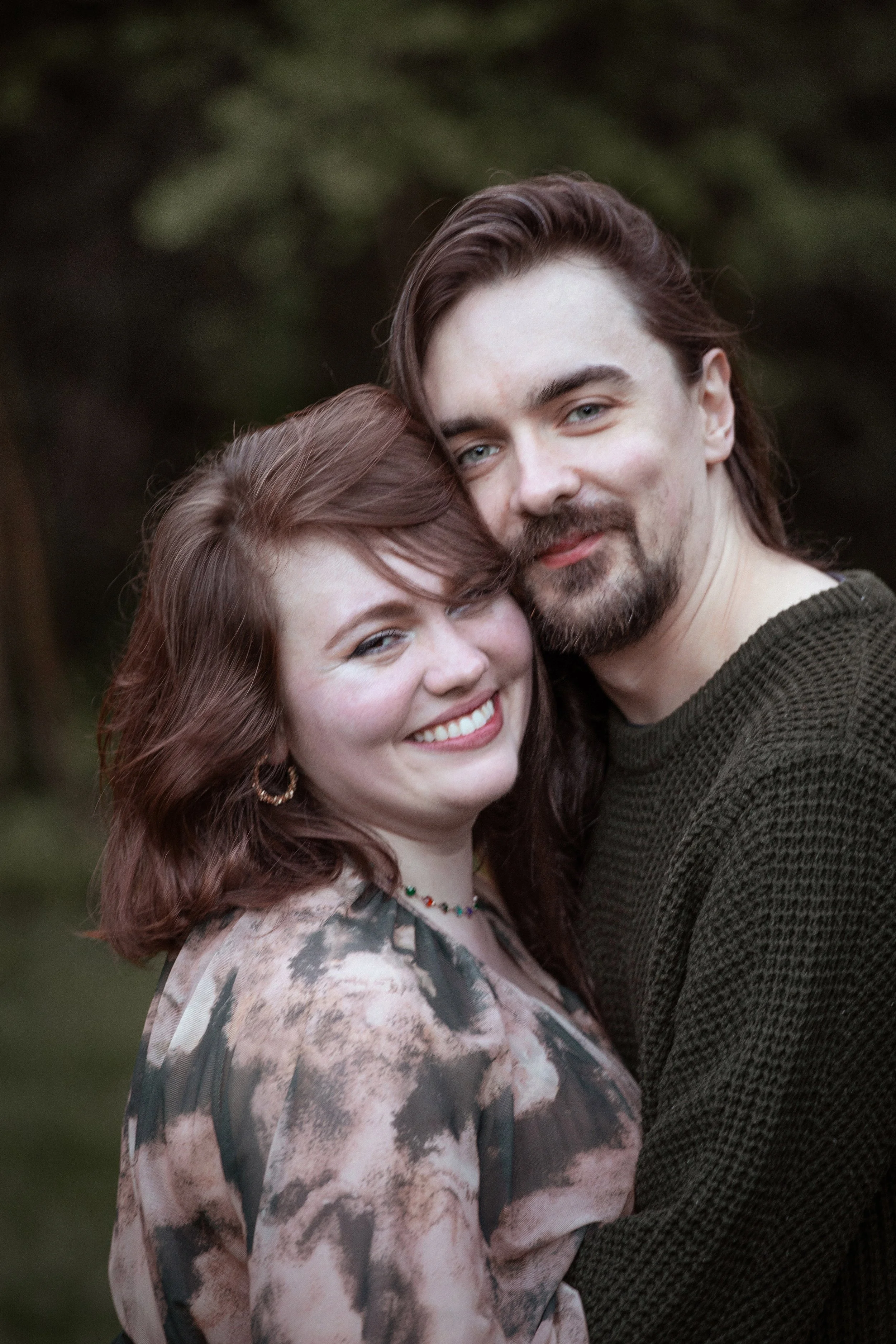 A smiling couple hugging outdoors, woman with shoulder-length red hair, wearing a patterned blouse and jewelry, man with long dark hair and a beard, wearing a dark sweater.