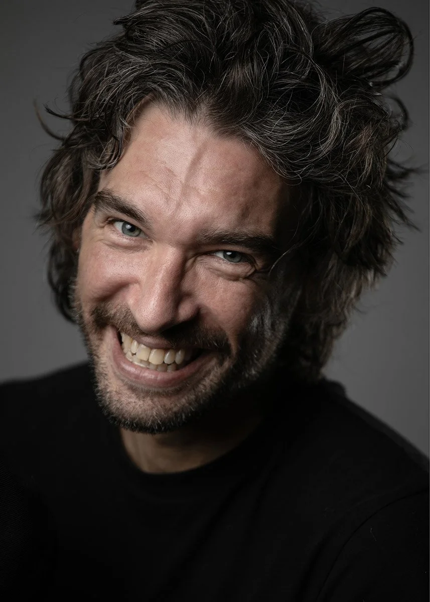 Close-up of a man with curly grayish-brown hair and a beard, smiling and showing his teeth, wearing a black shirt against a dark background.