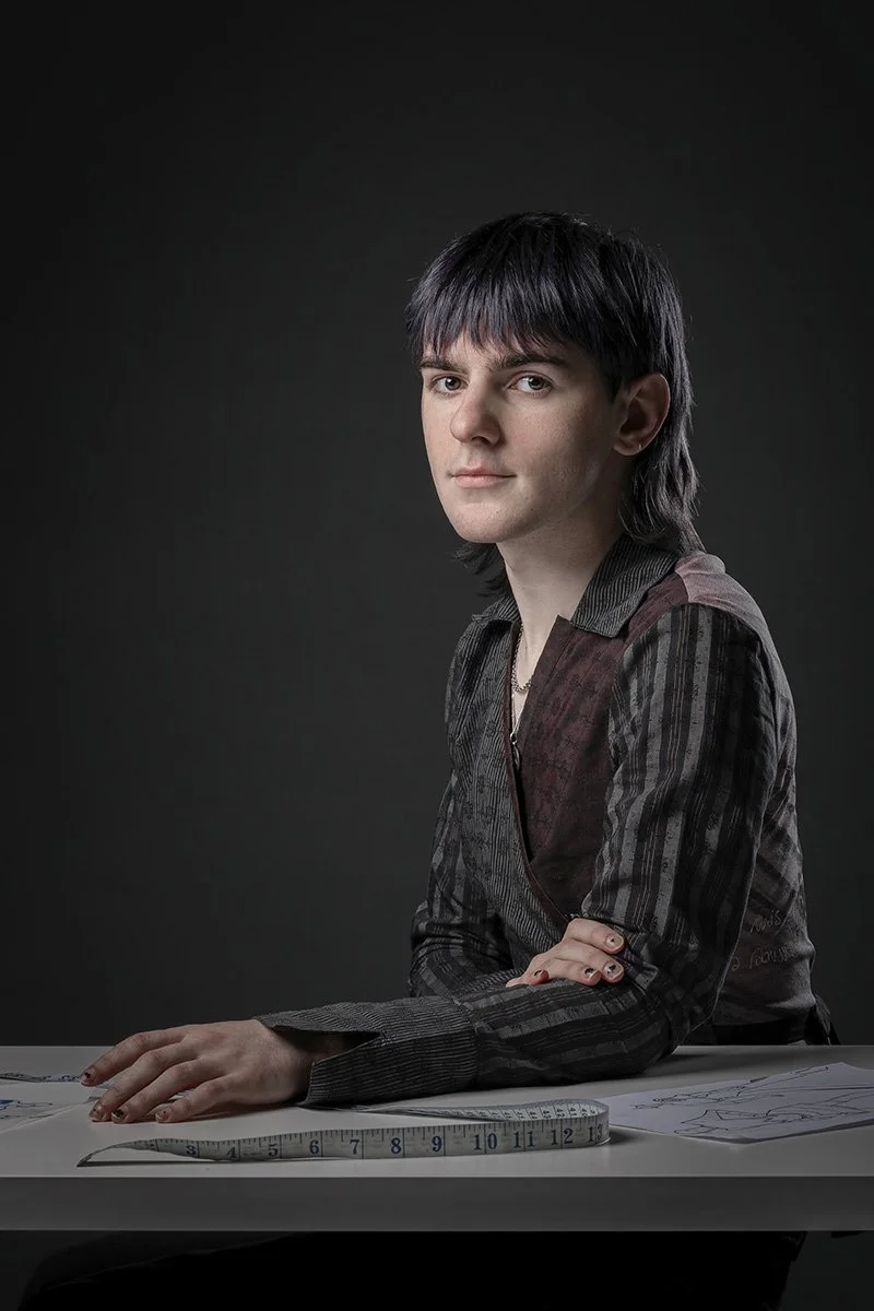 A young man with black hair and fair skin sitting at a table with a measuring tape and sketches, wearing a patterned shirt, against a dark background.
