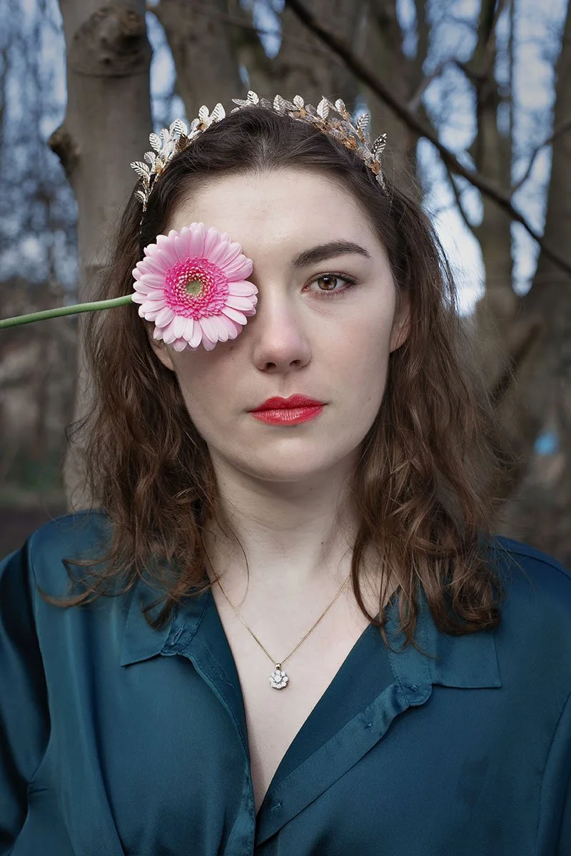 A woman with wavy brown hair, wearing a silver leaf crown, holds a pink flower over her left eye. She is wearing a dark teal blouse and a necklace with a circular pendant, standing outdoors in front of a tree with bare branches.