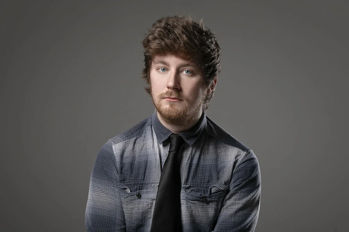A young man with brown, curly hair and a beard, wearing a gray plaid shirt and black tie, looking at the camera against a plain gray background.