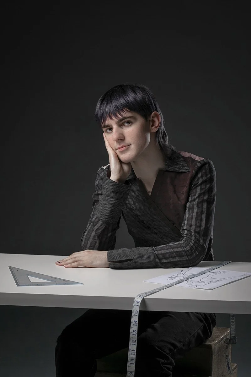 A person sitting at a white table with a tape measure, triangle ruler, and sketches, resting their head on their hand, with a thoughtful expression.