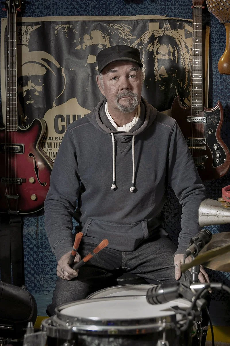 A man sitting behind a drum set holding drumsticks in a music studio, with guitars hanging on the wall behind him.
