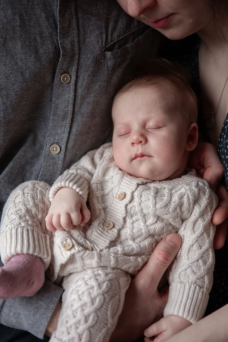 A close-up of a sleeping baby wearing a knitted cream-colored sweater and pants, cradled in someone's arms, with part of an adult's face and hand visible.
