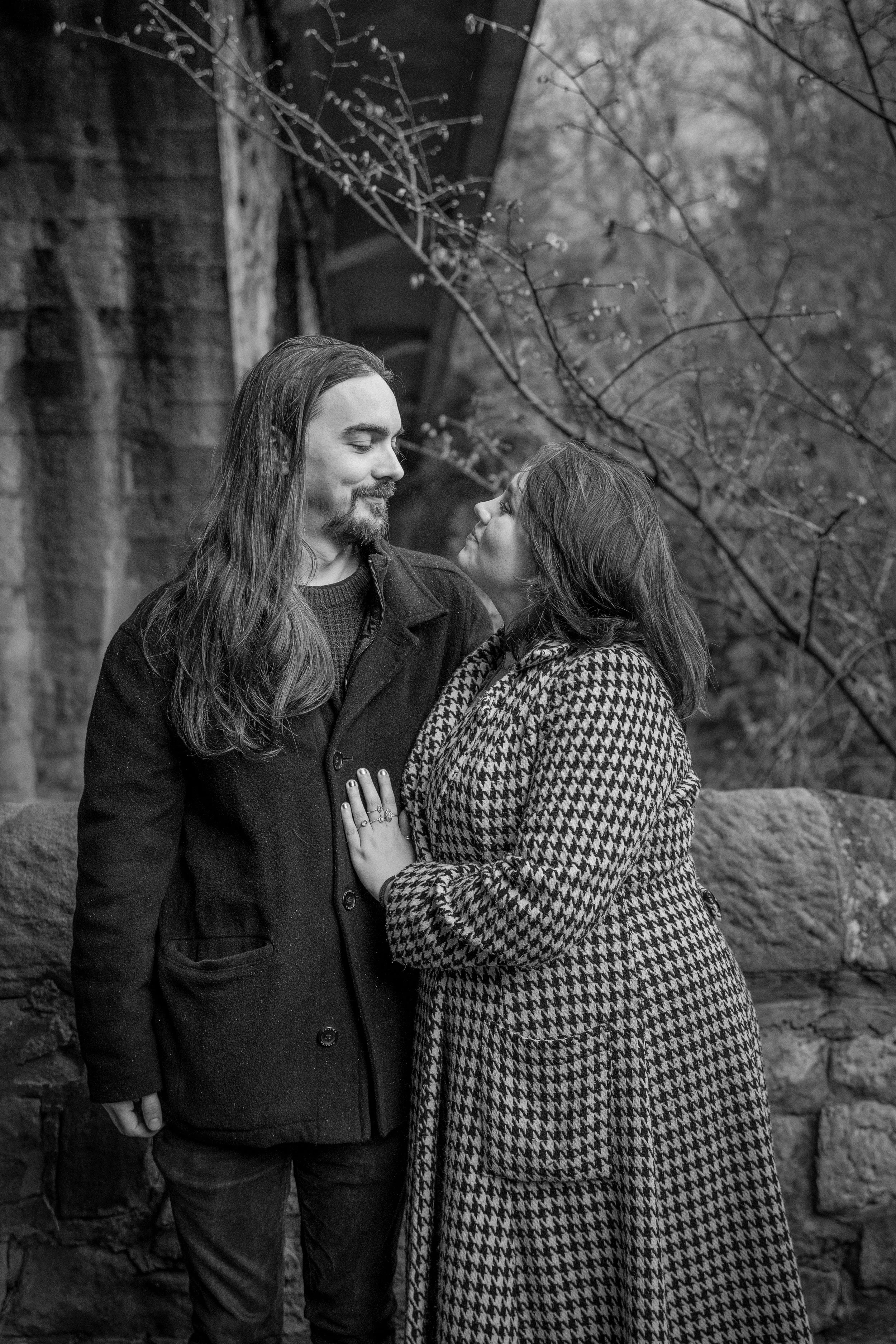 A black and white photo of a couple looking at each other affectionately outdoors, with trees and a stone wall in the background.