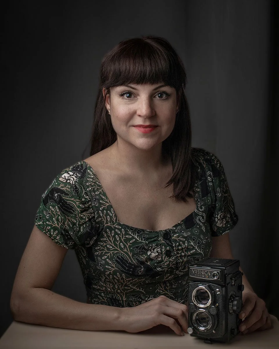 A woman with dark brown hair and bangs, wearing a patterned green dress, sitting at a table with a vintage twin-lens reflex camera in front of her against a dark background.