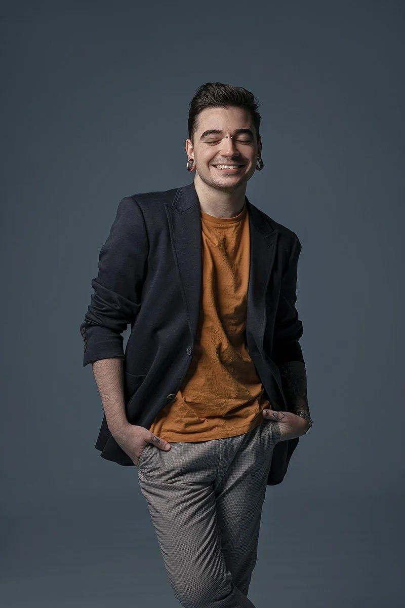 Smiling young man with dark hair, earrings, and tattoos wearing a black blazer over an orange t-shirt and gray pants, standing with hands in pockets against a solid dark background.