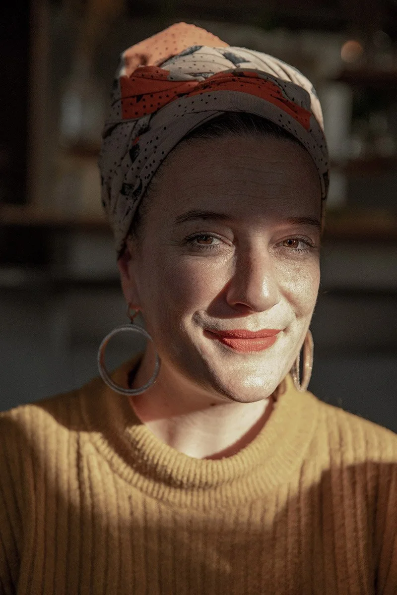 A woman smiling, wearing a colorful headscarf and large hoop earrings, in warm lighting.
