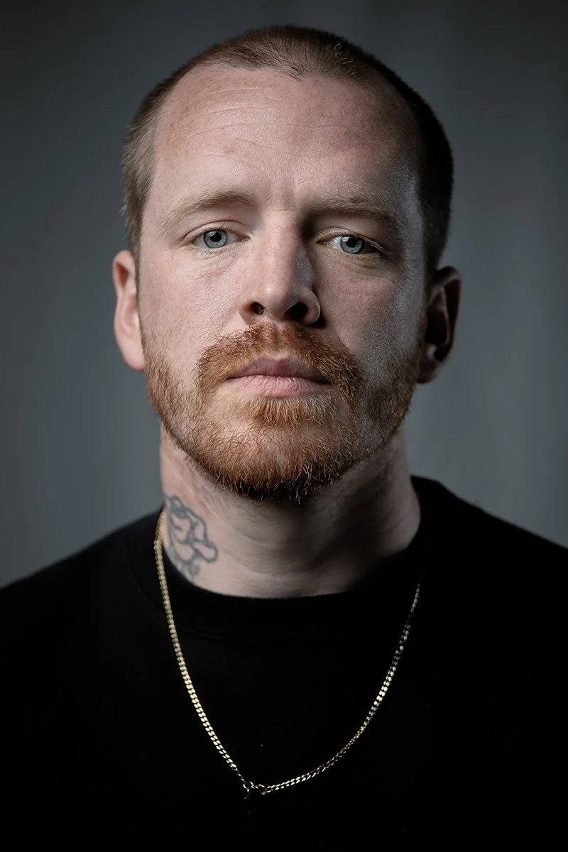 Close-up portrait of a man with light skin, short hair, a reddish beard, blue eyes, a tattoo on his neck, wearing a black shirt and a gold chain necklace.