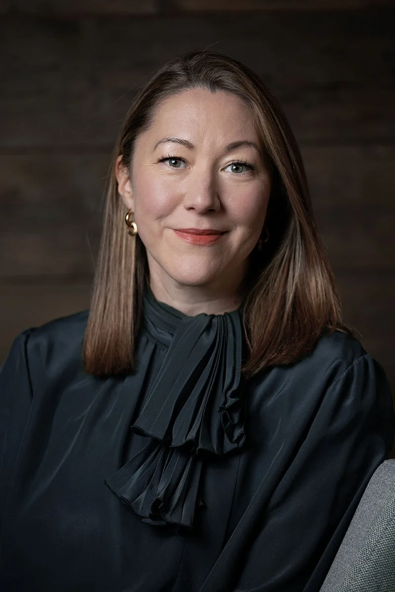 A woman with shoulder-length brown hair, blue eyes, and light makeup, wearing a dark blouse with a ruffled bow neck and gold earrings, smiling in front of a dark wooden background.