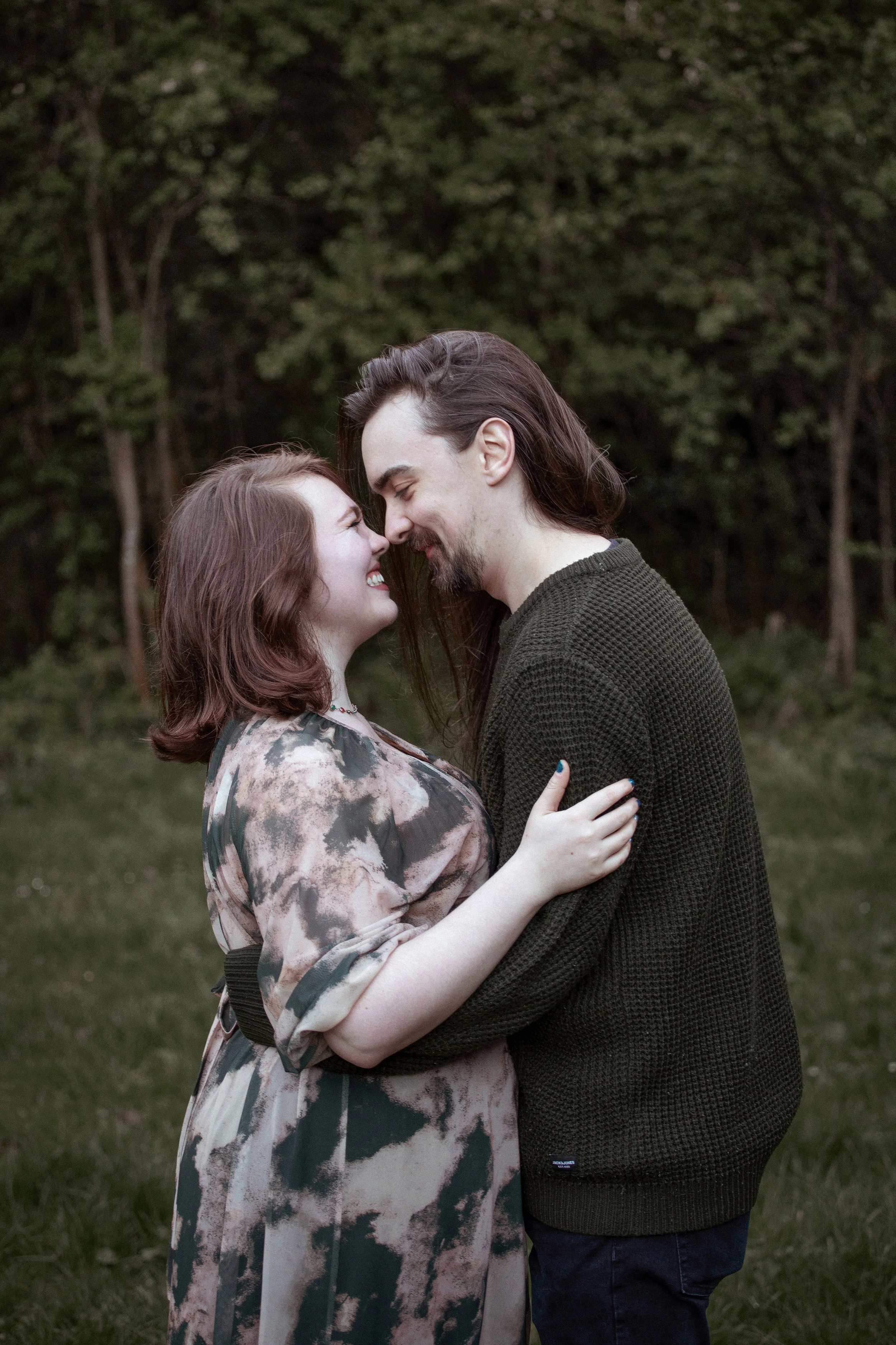 A couple with their foreheads touching and smiling in a park with trees in the background.