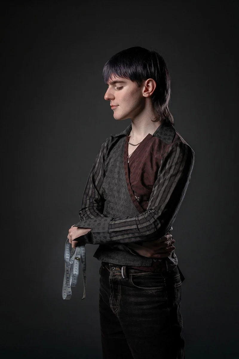 A young man with black hair and fair skin posing with arms crossed, holding a measuring tape in his right hand, against a plain dark background.