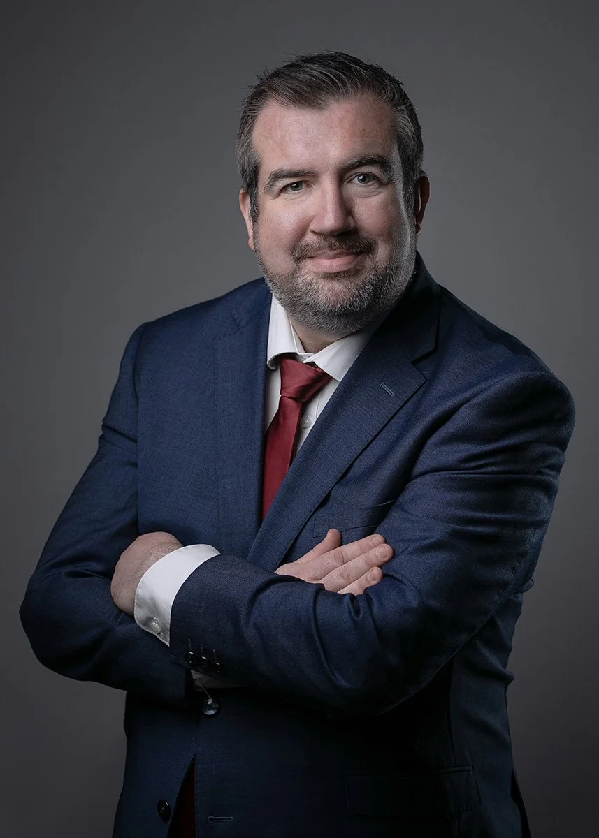 A professional man with dark hair and a beard, wearing a navy suit, white shirt, and red tie, standing with arms crossed against a gray background.