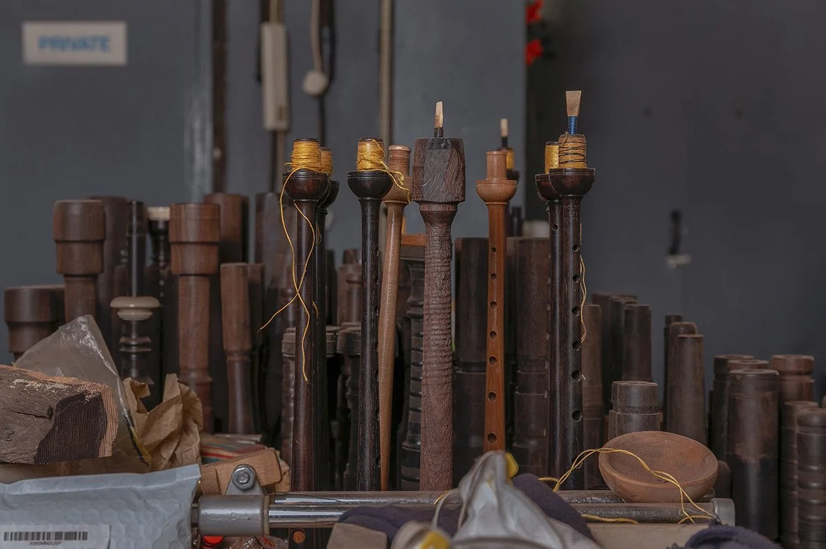A collection of various wooden parts for musical instruments, likely recorders or similar wind instruments, including mouthpieces and body sections, placed on shelves in a workshop.