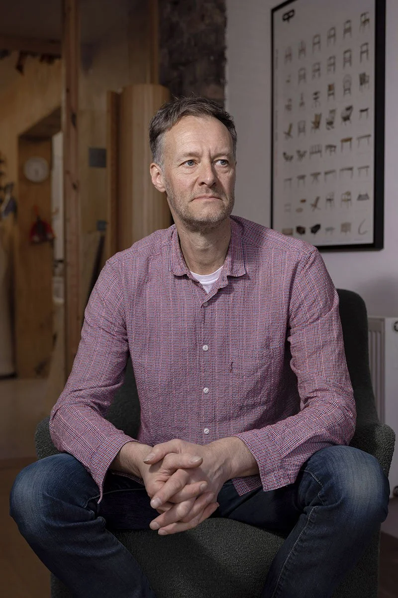 A man sitting in a chair in a room with wooden beams and a display of various tools or objects on the wall behind him.
