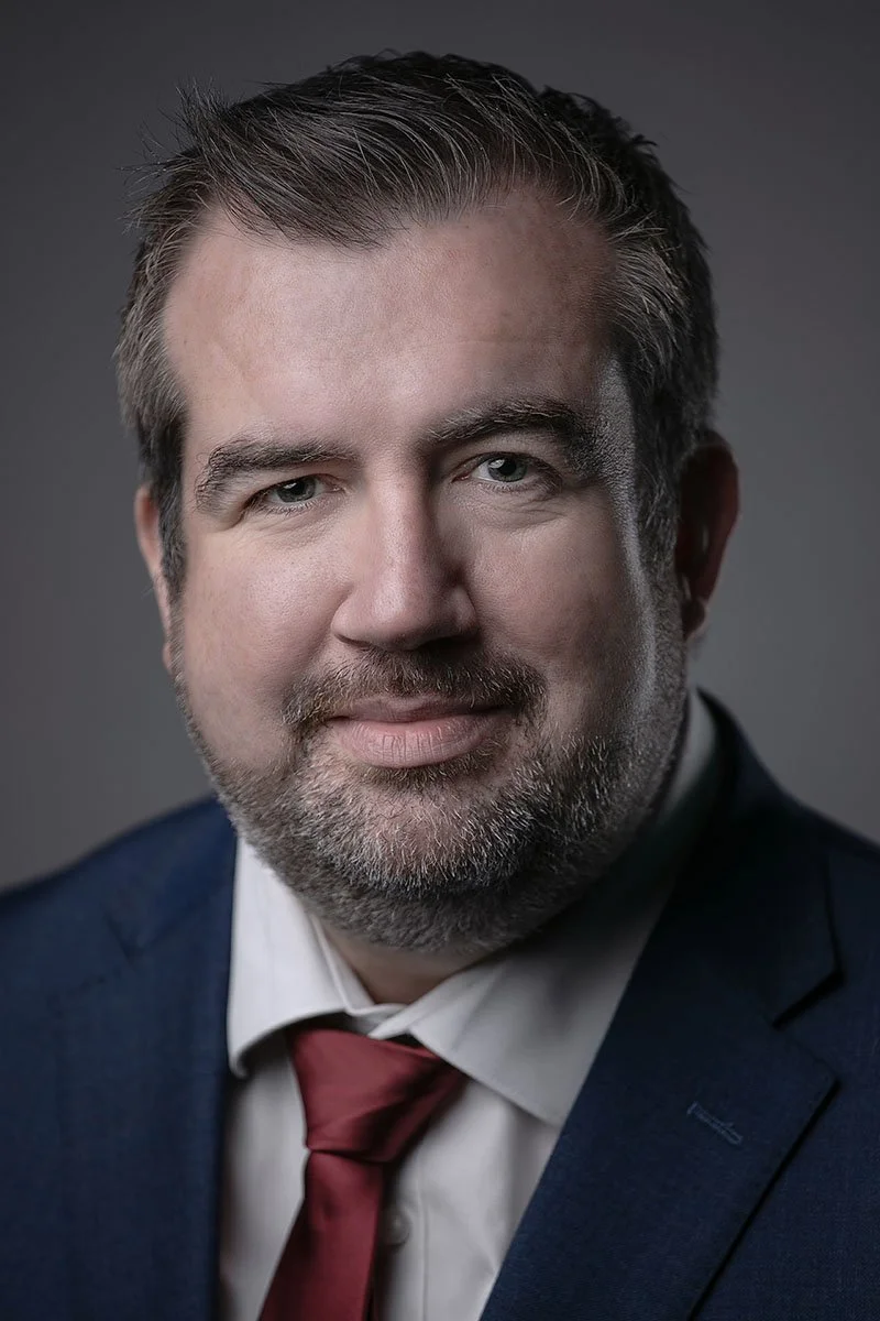 Headshot of a man with short dark hair, light beard, wearing a dark suit, white shirt, and red tie against a gray background.