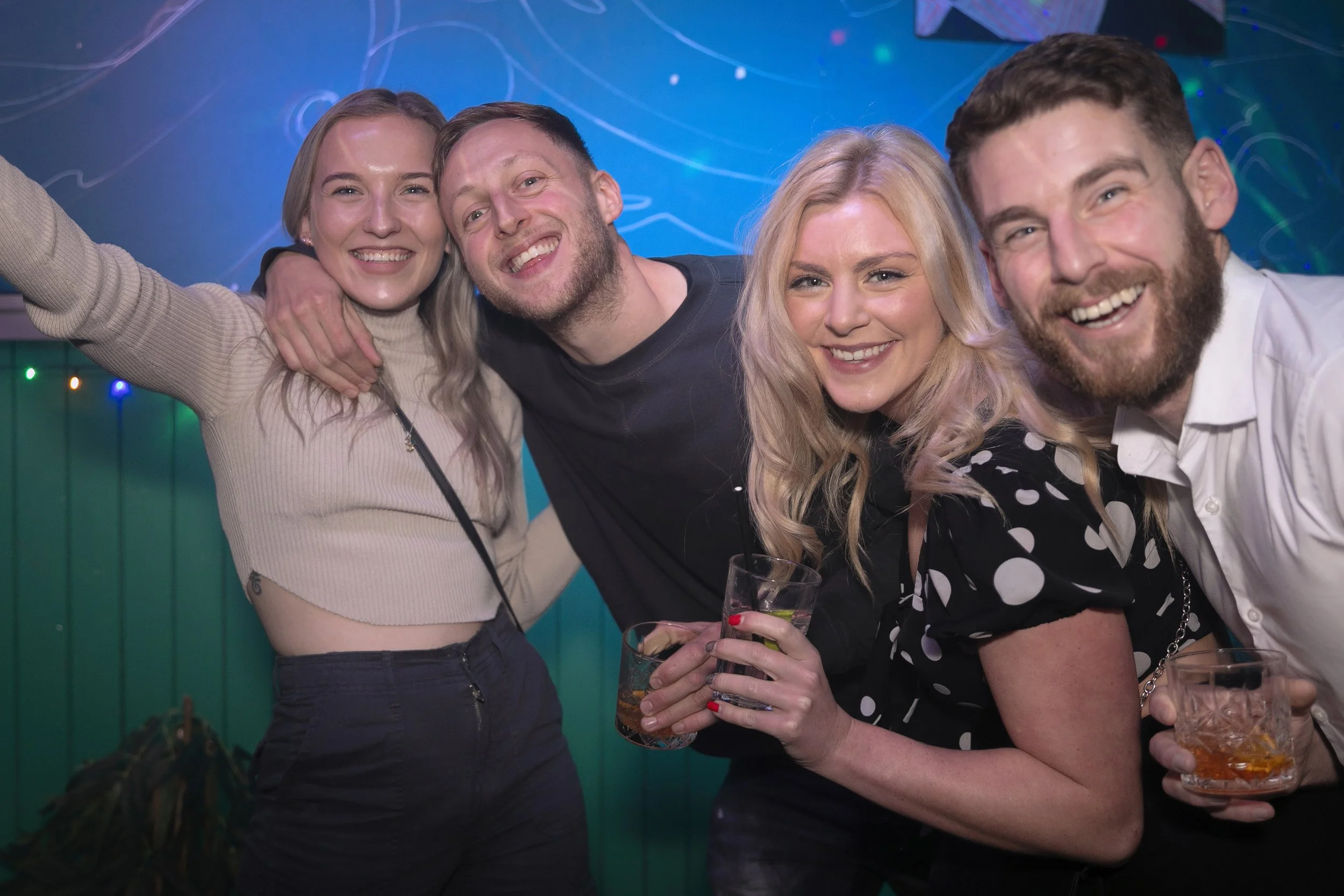 Four friends smiling and enjoying drinks at a party or bar, posing closely together for a group photo.