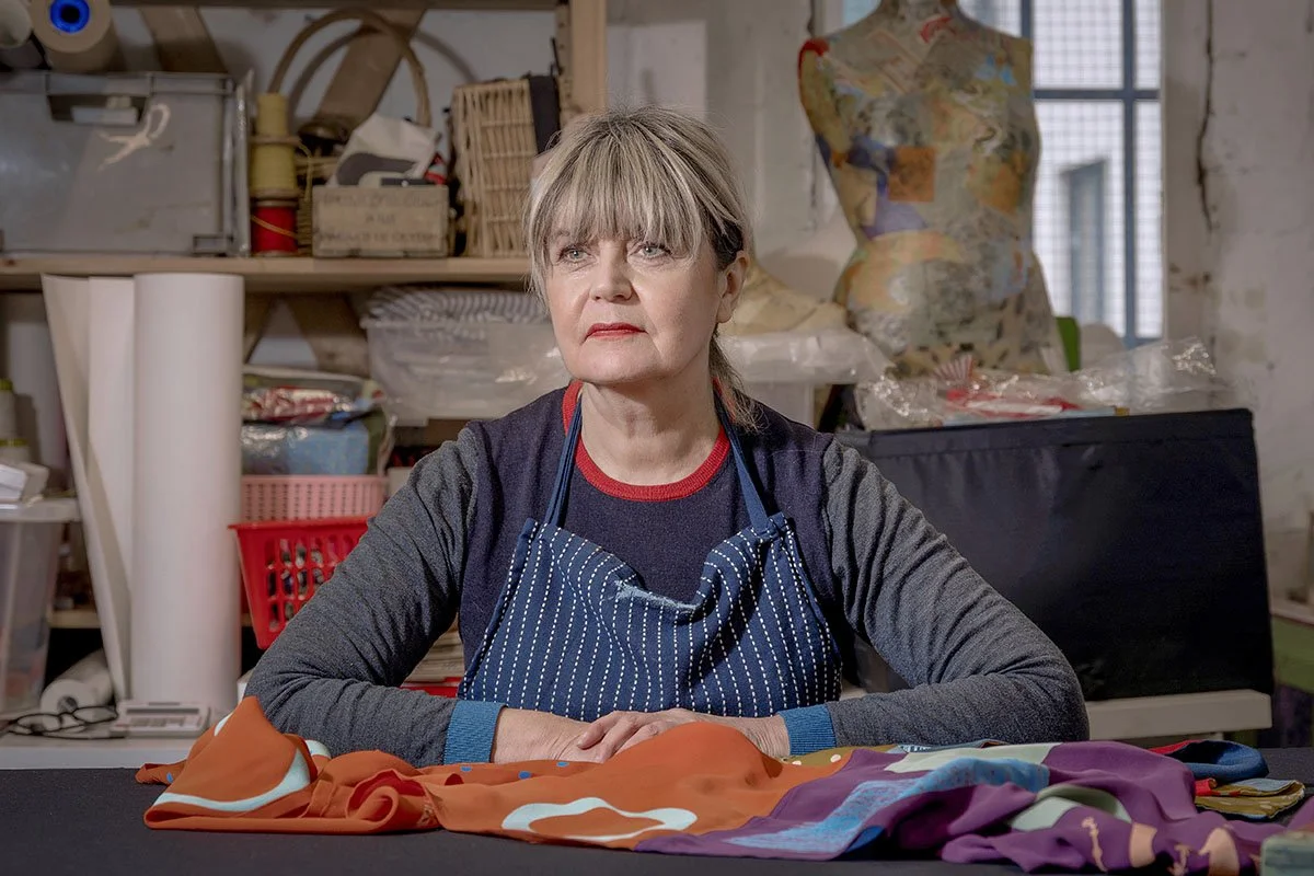 A woman with short blonde hair and a striped apron sitting at a worktable in a cluttered craft or sewing room.