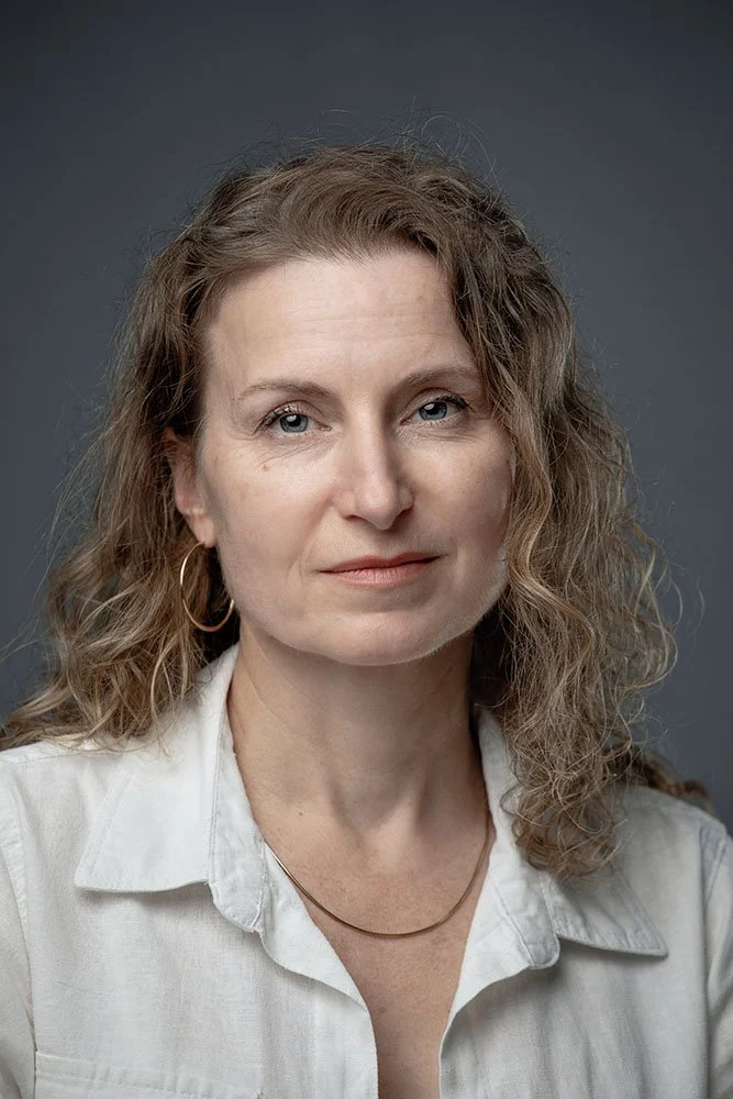 Portrait of a woman with wavy brown hair, blue eyes, wearing a white collared shirt, hoop earrings, and a simple necklace against a gray background.