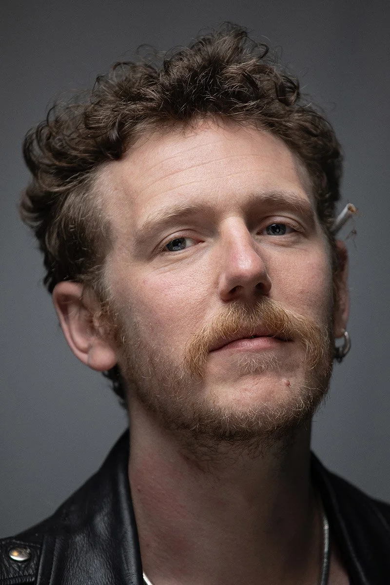 A close-up portrait of a young man with curly hair and a mustache, wearing a leather jacket and earrings, against a plain backdrop.