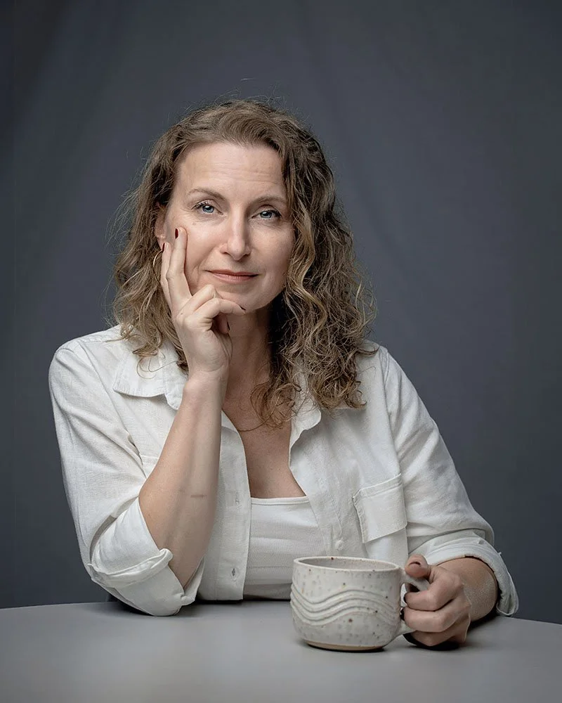 A woman with curly hair sitting at a table, resting her chin on her hand, holding a mug in her other hand, with a neutral gray background.