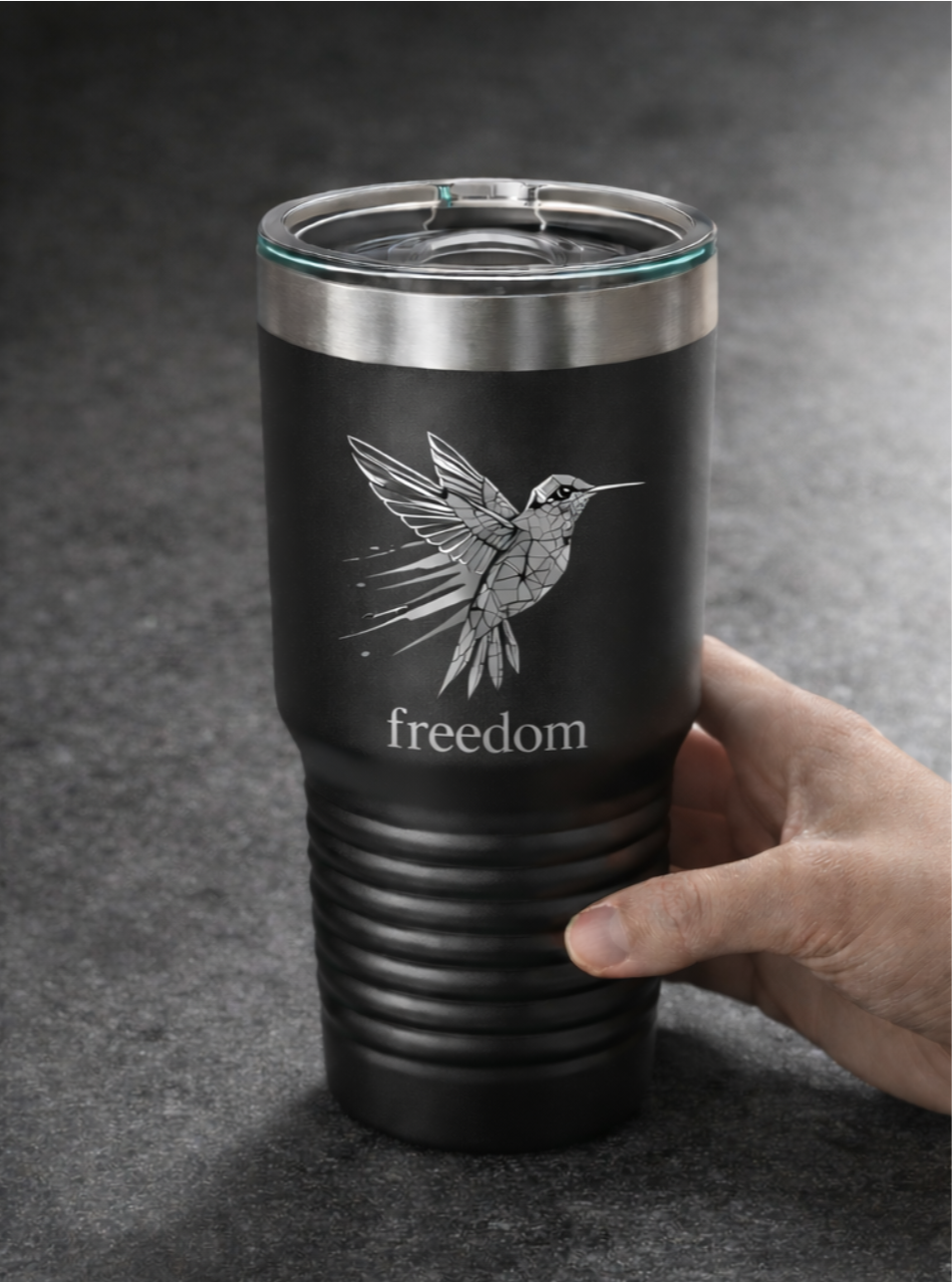 A black travel tumbler with a stainless steel rim and a clear lid, featuring a geometric illustration of a hummingbird and the word "freedom" written below.