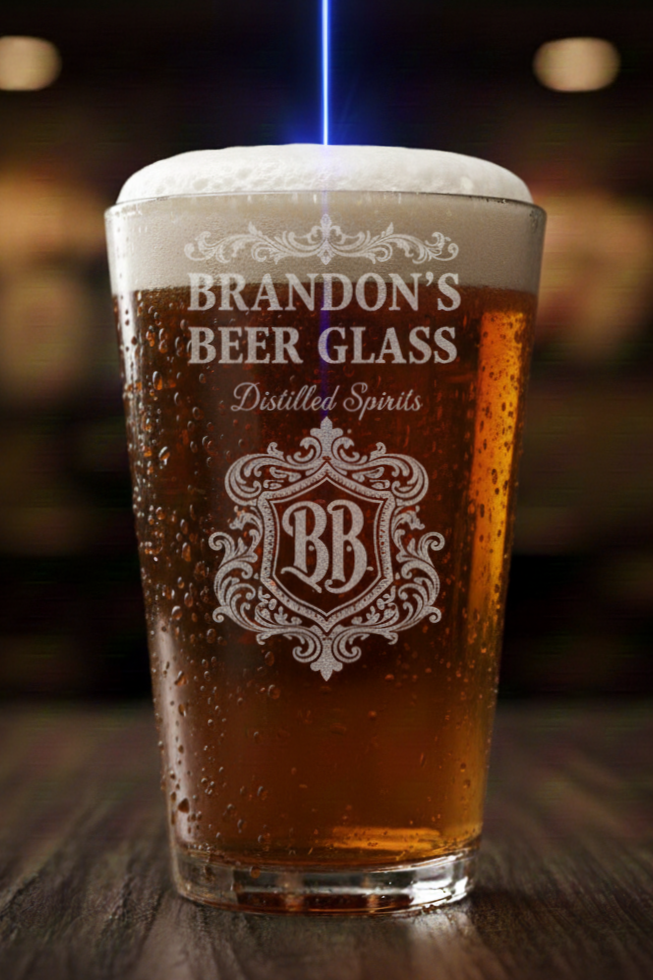 A pint glass filled with beer, topped with foam, with a glass or bottle in the background. The glass has a logo and text that reads 'Brandon's Beer Glass' and 'Distilled Spirits'.