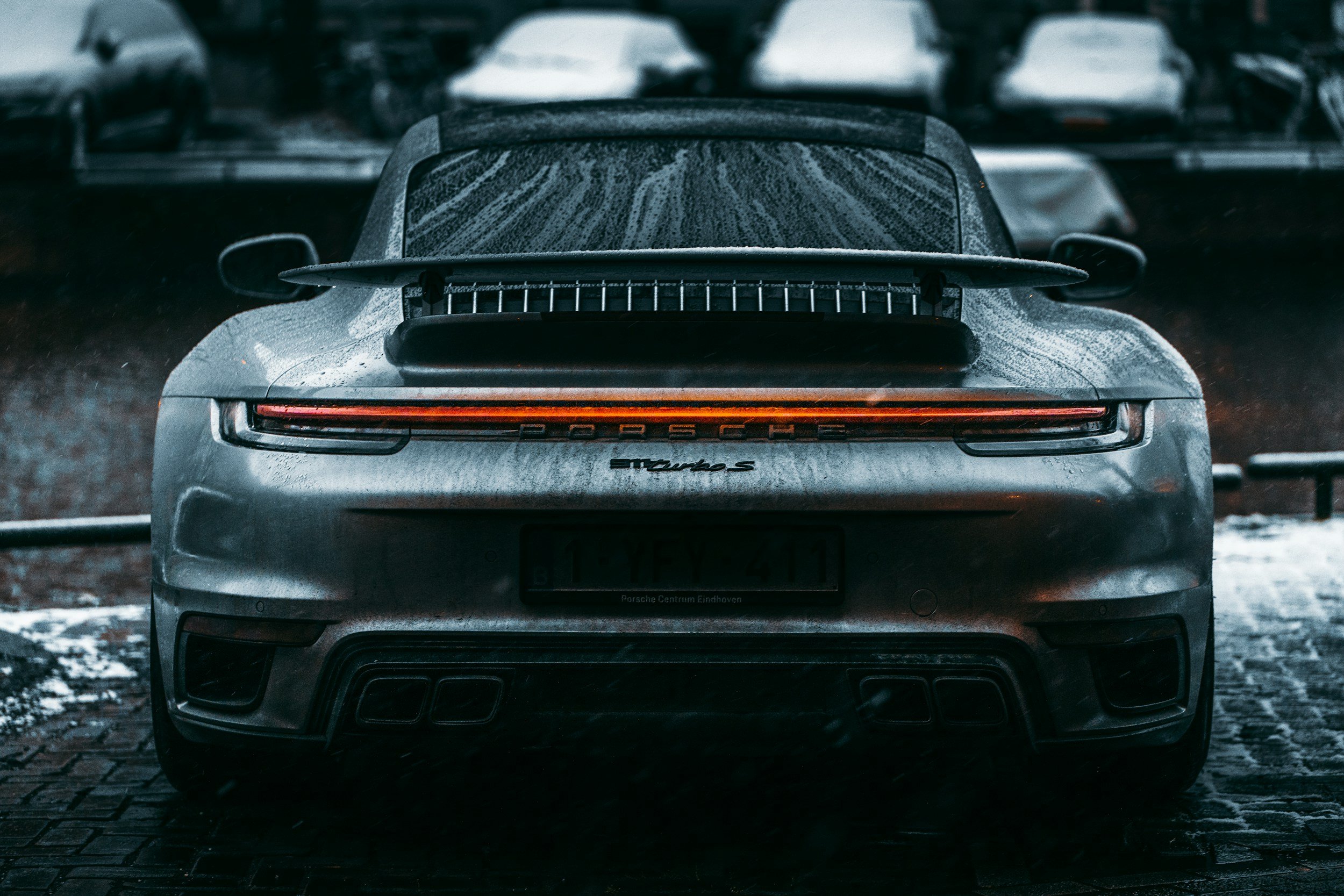 Rear view of a silver Porsche 911 Turbo S parked on a wet street, with water droplets on the car and blurred cars in the background.