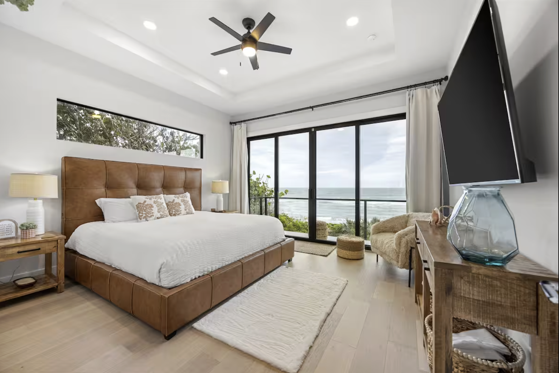 A bedroom with a large bed, white bedding, and beige decorative pillows, a wooden nightstand with a lamp on each side, a white area rug, a sliding glass door leading to a balcony with an ocean view, a beige armchair with a throw blanket, a wooden desk with a glass vase and TV on top, and a ceiling fan.
