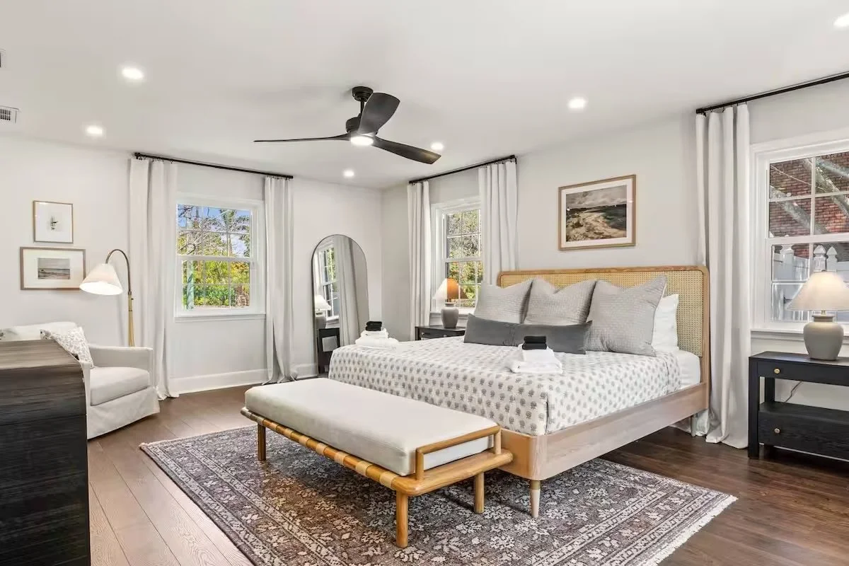 A bedroom with a large bed, white bedding, and beige decorative pillows, a wooden nightstand with a lamp on each side, a white area rug, a sliding glass door leading to a balcony with an ocean view, a beige armchair with a throw blanket, a wooden desk with a glass vase and TV on top, and a ceiling fan.