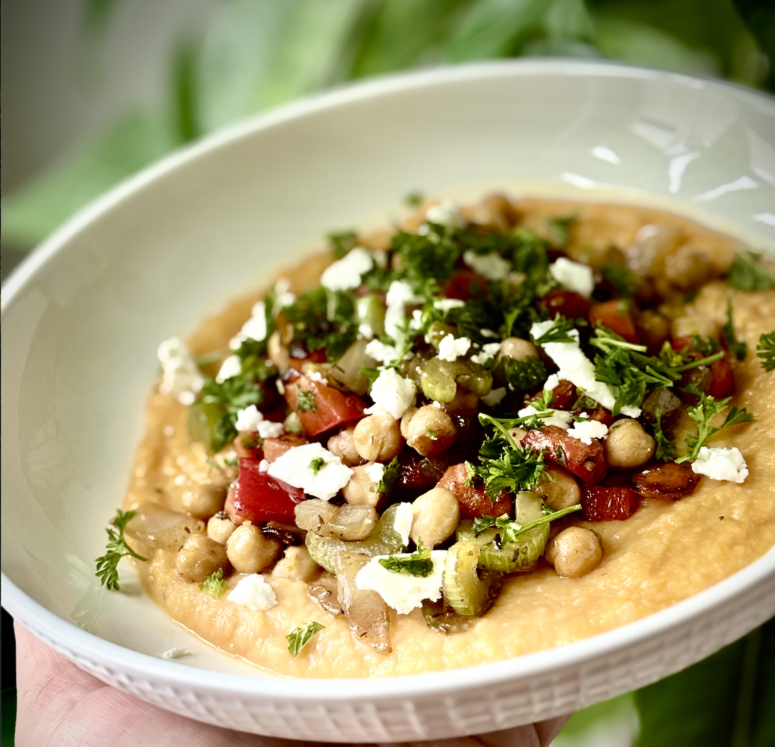 A bowl of creamy yellow dish topped with chopped tomatoes, chickpeas, chopped onions, crumbled cheese, chopped parsley, and cilantro.