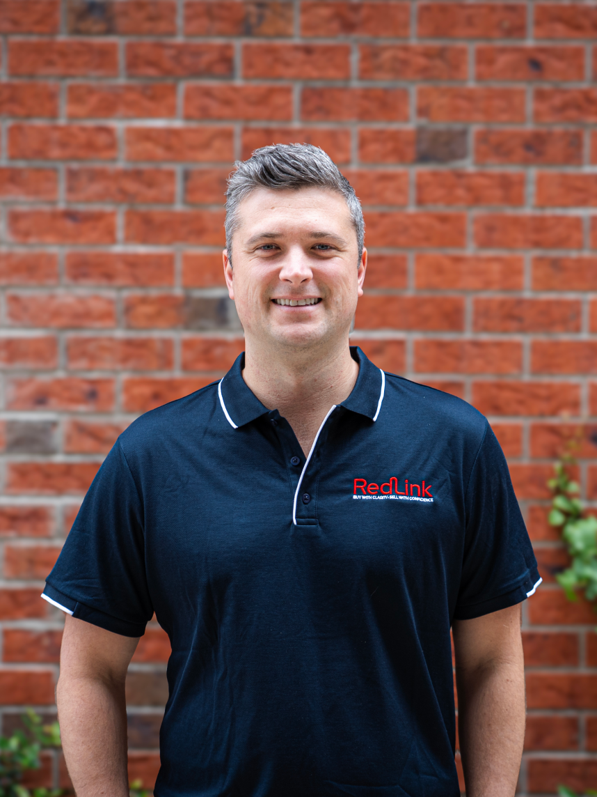 A man with curly dark hair, beard, and glasses, wearing a black sweater, looking thoughtfully to the side indoors with a blurred plant in the background.