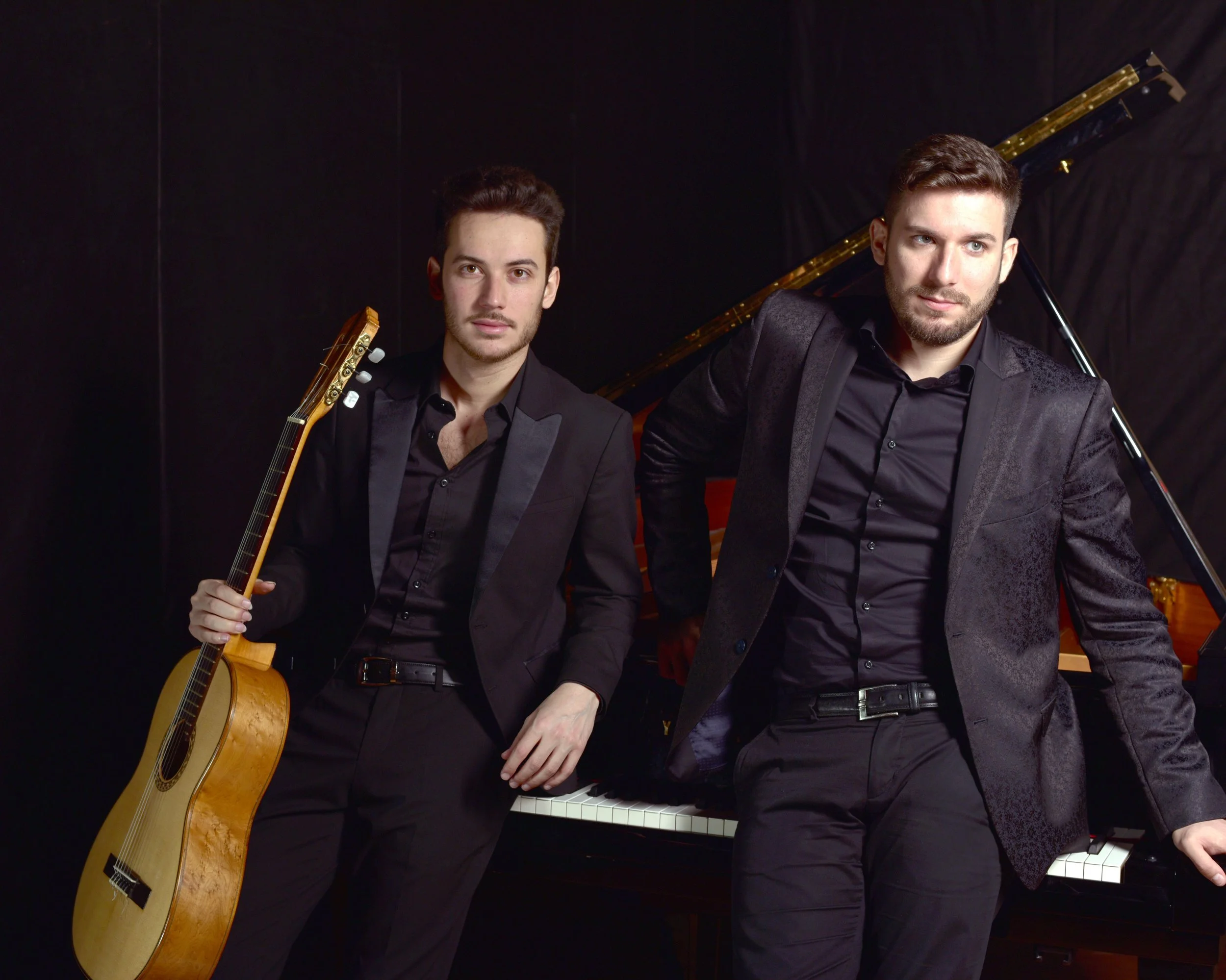 Due uomini in abiti neri con una chitarra classica e un pianoforte nero con copertura in velluto, in un ambiente scuro.