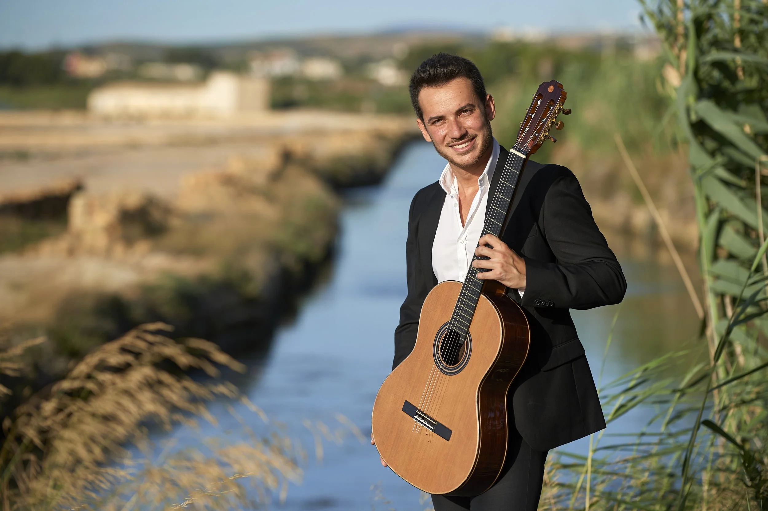 Uomo sorridente con abito nero e camicia bianca, che tiene una chitarra acustica, in riva a un fiume con paesaggio naturale e cielo blu.