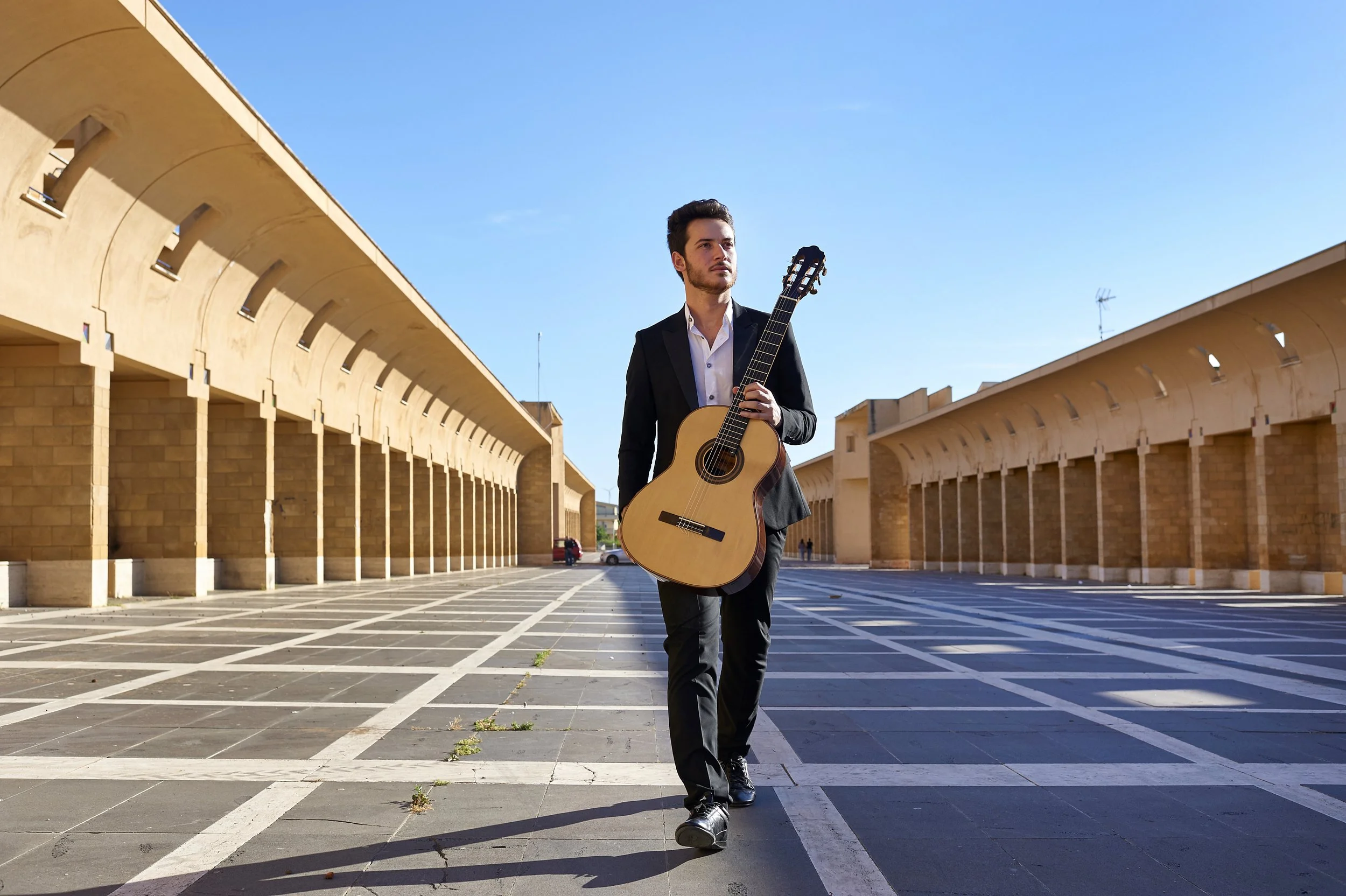 Un uomo in abito nero cammina con una chitarra classica in mano attraverso una piazza vuota sotto un cielo azzurro.