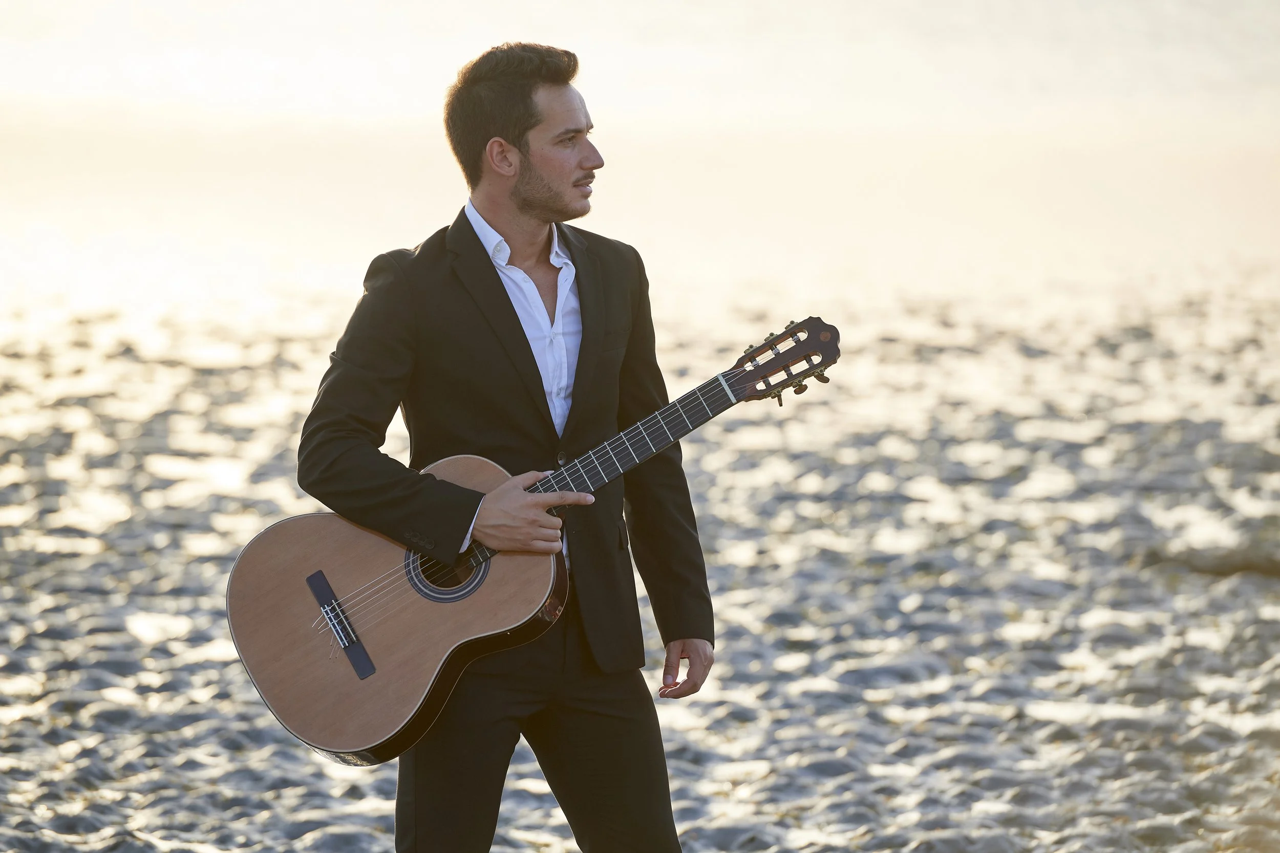 Uomo in giacca nera con chitarra classica al tramonto sulla spiaggia