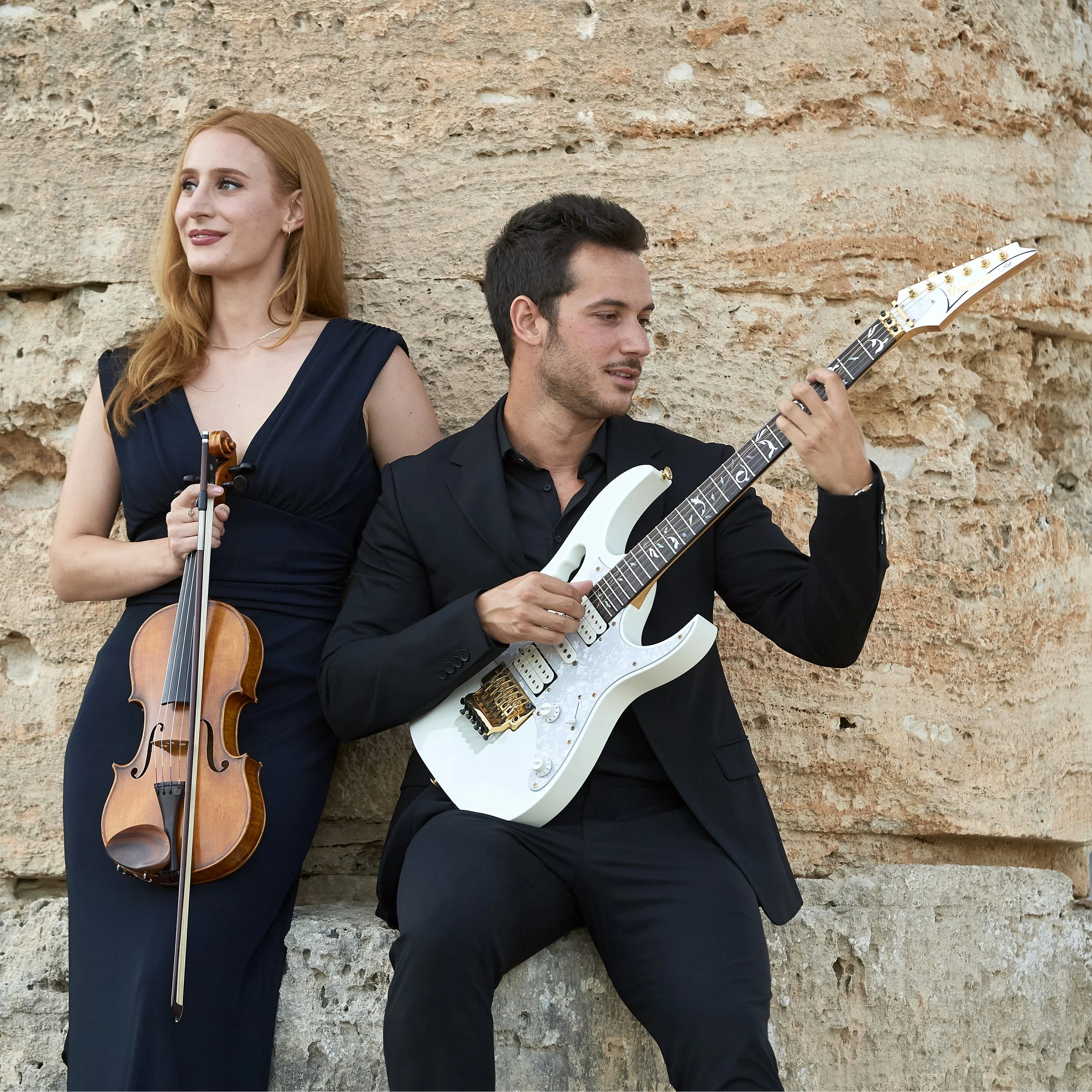 Due musicisti, una donna con un violino e un uomo con una chitarra elettrica, si trovano all'aperto vicino a un muro di pietra