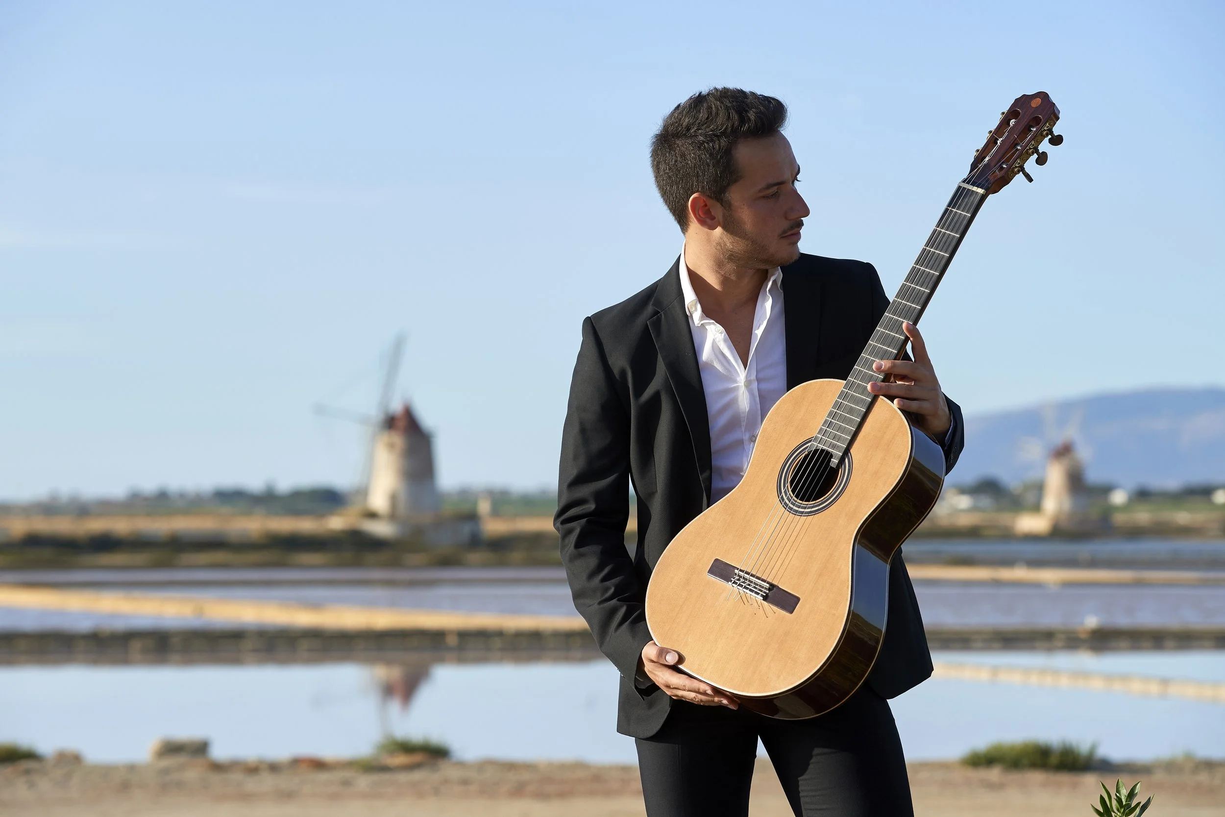 Un uomo in abito nero suona una chitarra acustica all'aperto con un paesaggio di mulini a vento in lontananza.