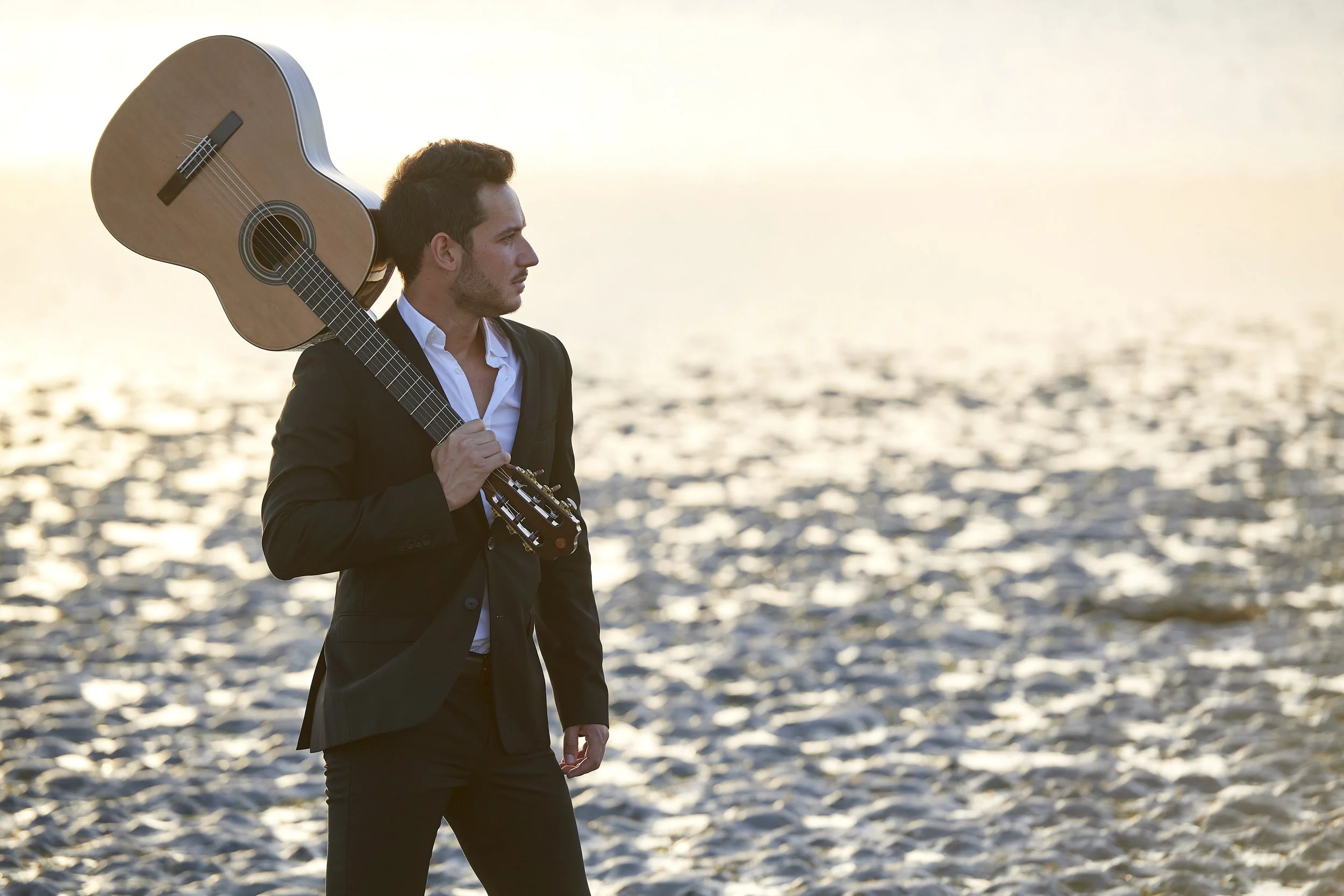 Un uomo in giacca nera e camicia bianca tiene una chitarra sotto il braccio, camminando sul bagnasciuga durante il tramonto.