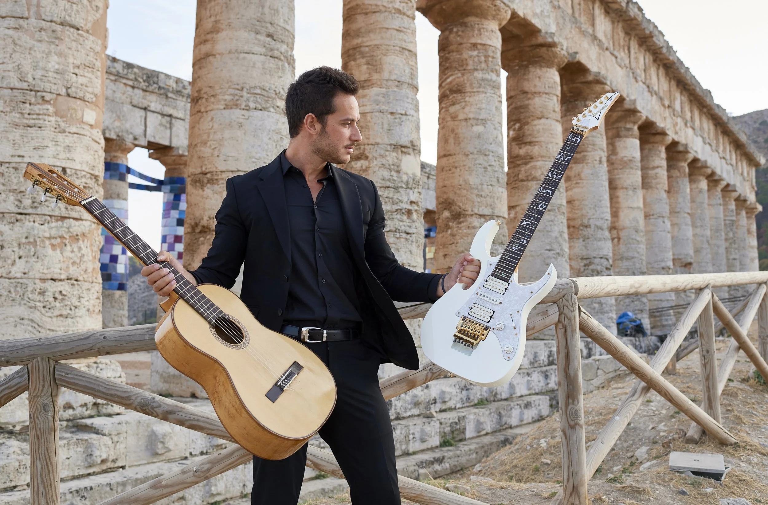Uomo in abiti eleganti con due chitarre, una acustica e una elettrica, davanti a antiche colonne di pietra in un sito archeologico.