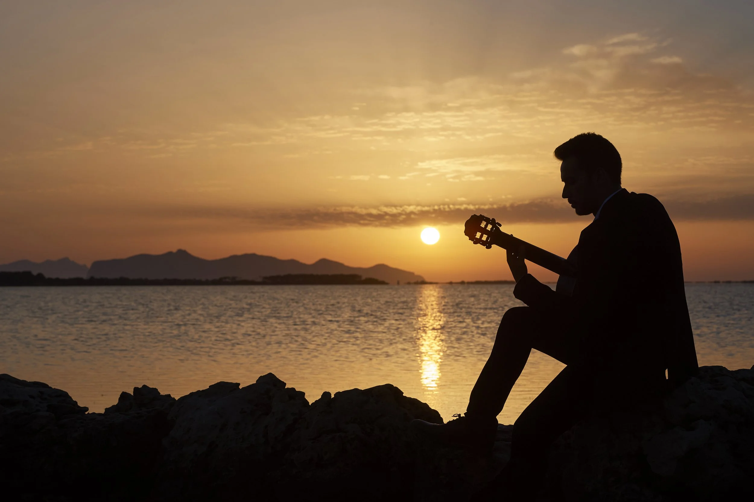 Siluetta di un uomo che suona la chitarra seduto su una roccia durante il tramonto, vista sul mare e montagne in lontananza.