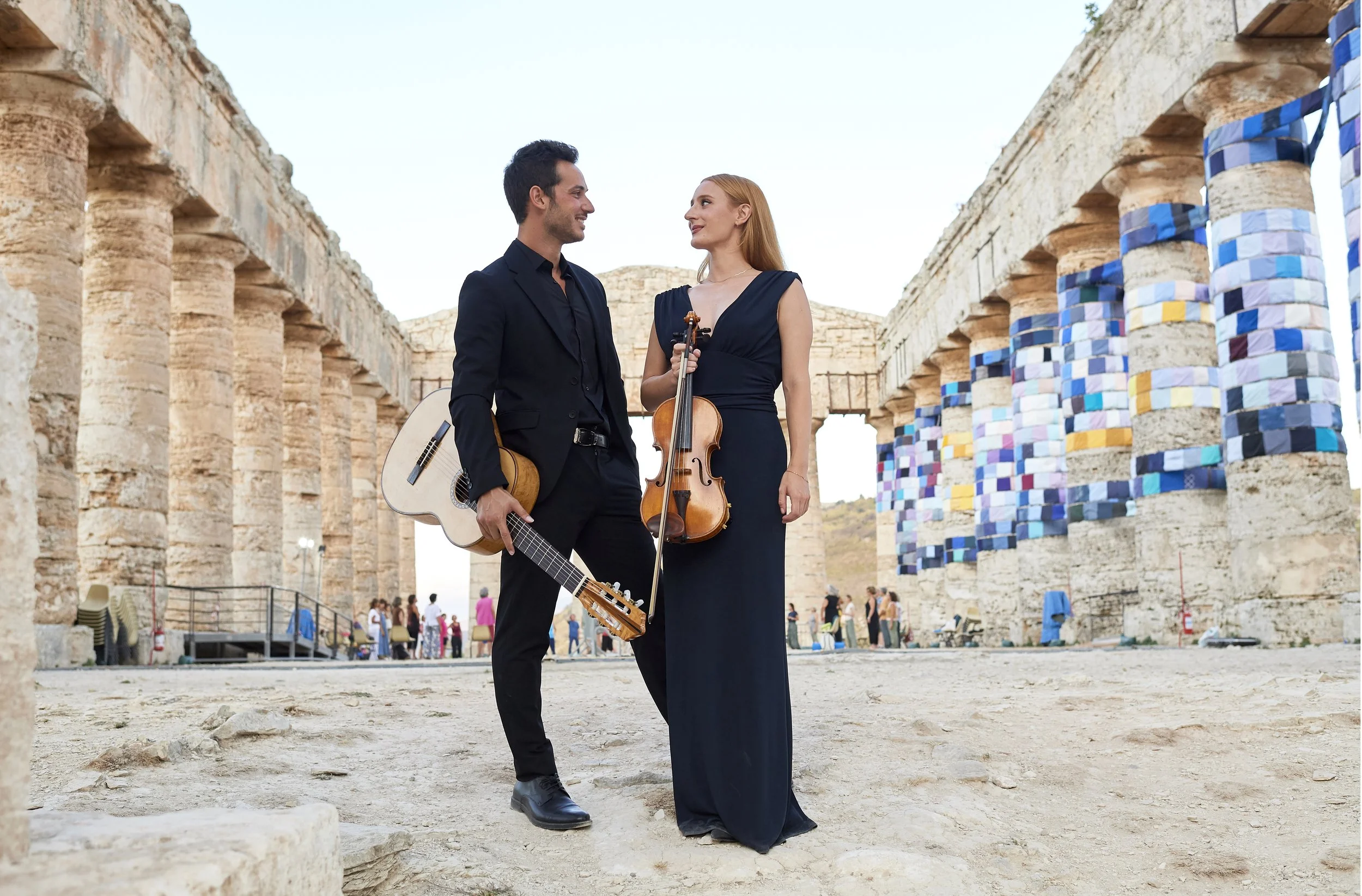 Un uomo e una donna vestiti elegantemente si incontrano e sorridono, ciascuno tiene uno strumento musicale, un canto e un violino, in un'area archeologica con colonne antiche decorate con tessere di mosaico colorate.