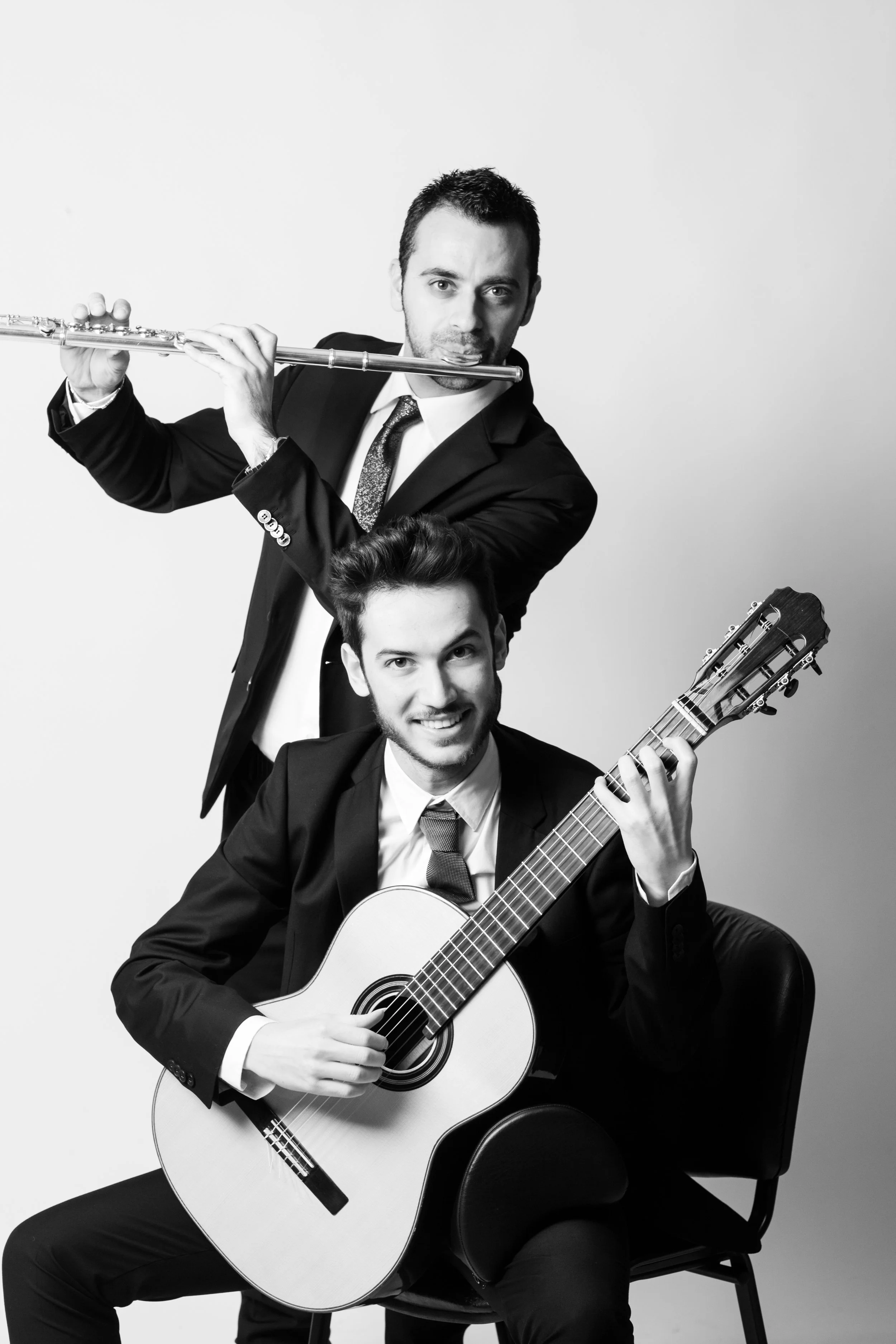 Due uomini in abiti eleganti, uno seduto con chitarra e uno in piedi con flauto, sorridono in uno studio con sfondo bianco.