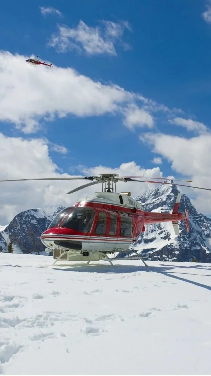 A red and white helicopter on snow with mountains in the background, and another helicopter flying in the sky above.